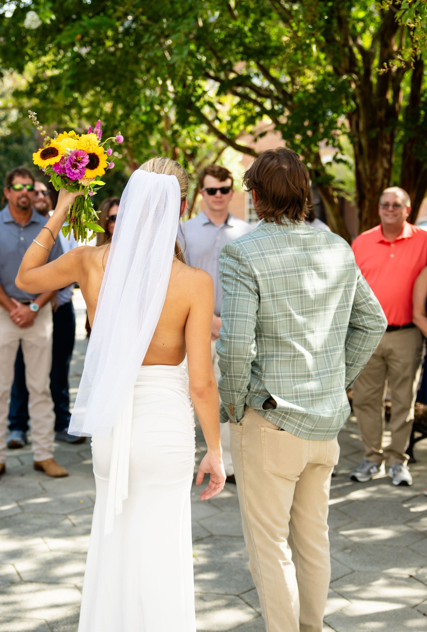 Back view of bride in white backless gown and veil holding vibrant bouquet with wedding guests.