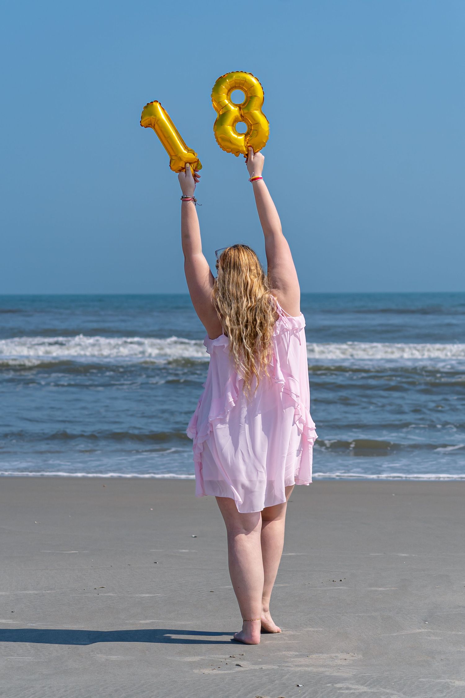 Young woman in pink dress holding gold number 18 balloons on beach, celebrating her 18th birthday by the ocean.