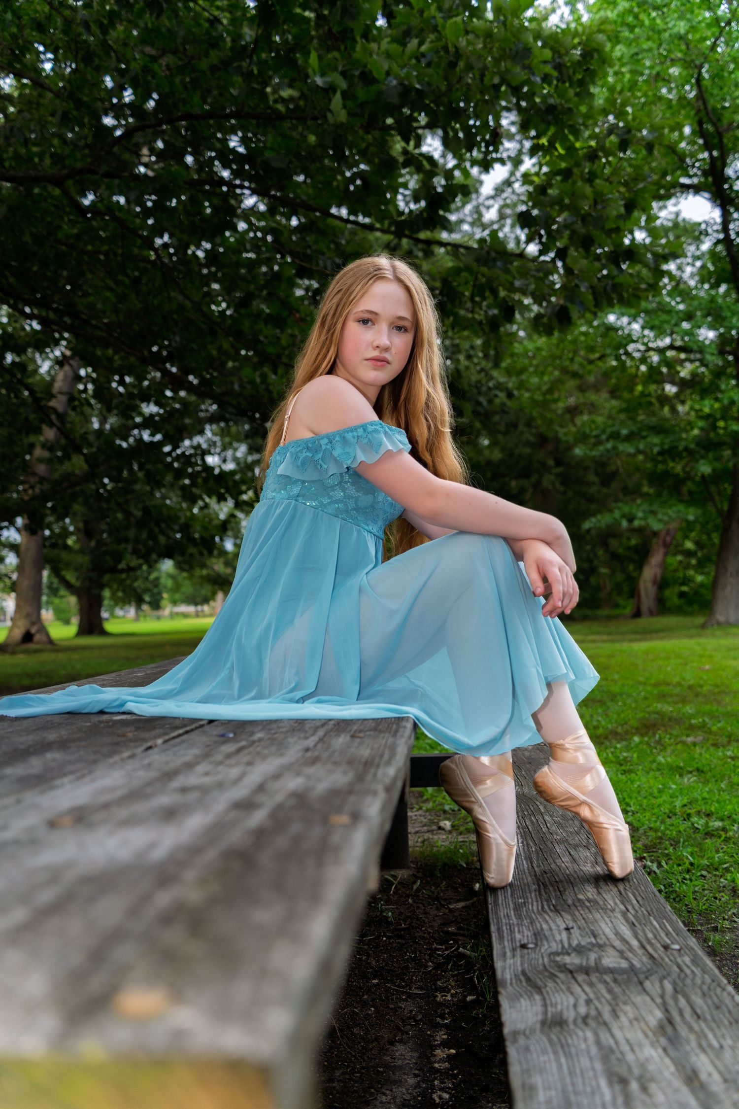 Ballet dancer in light blue dress rests gracefully on wooden platform in a serene garden setting.
