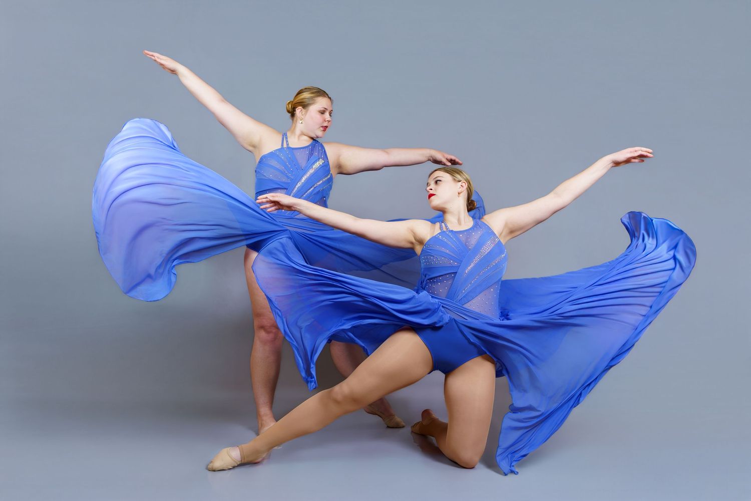 Two female dancers in flowing blue dresses perform an elegant contemporary duet against a gray studio background.