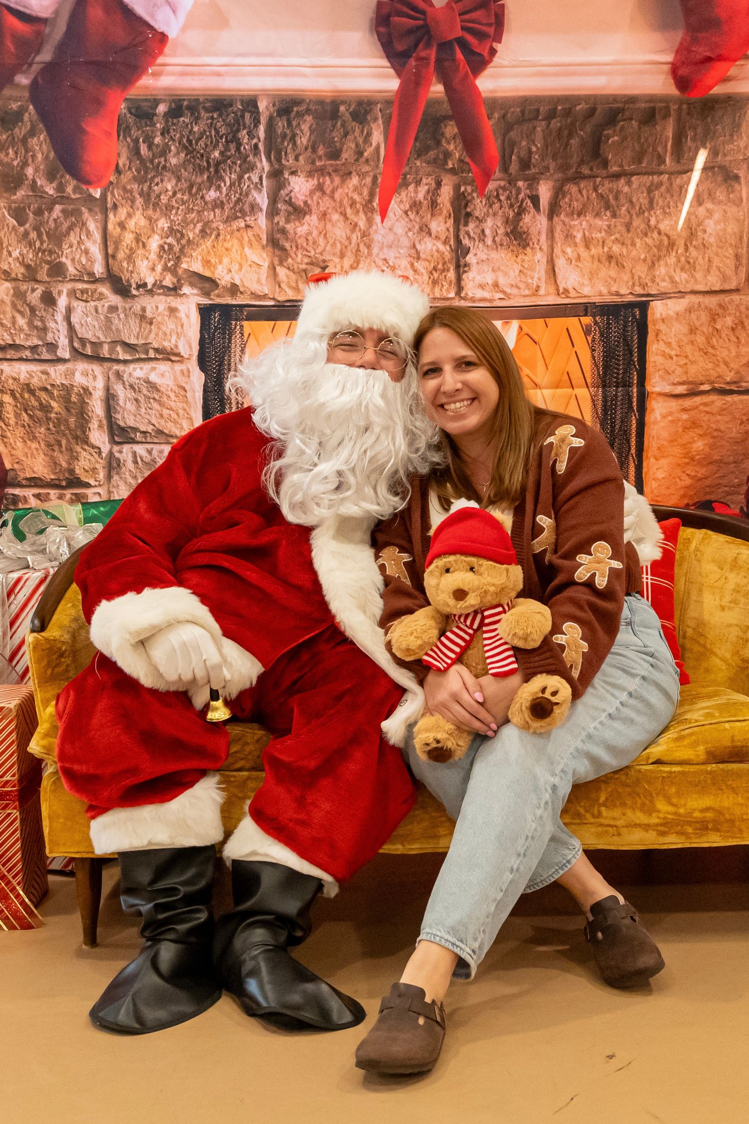 Santa and a visitor pose with a teddy bear on a yellow couch in front of a decorated stone fireplace.