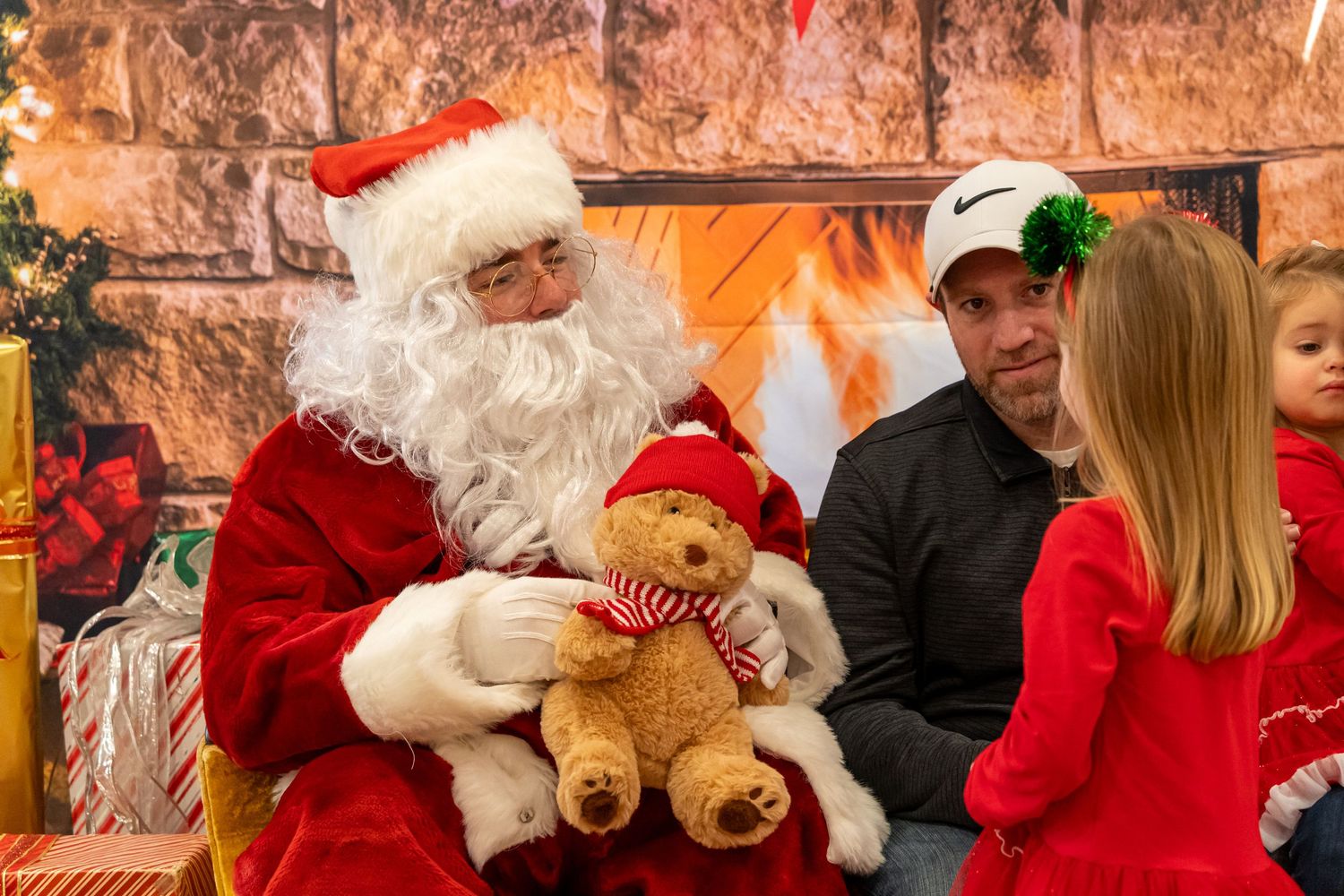 A holiday gathering with Santa and visitors sharing a moment with a teddy bear by a decorated fireplace.