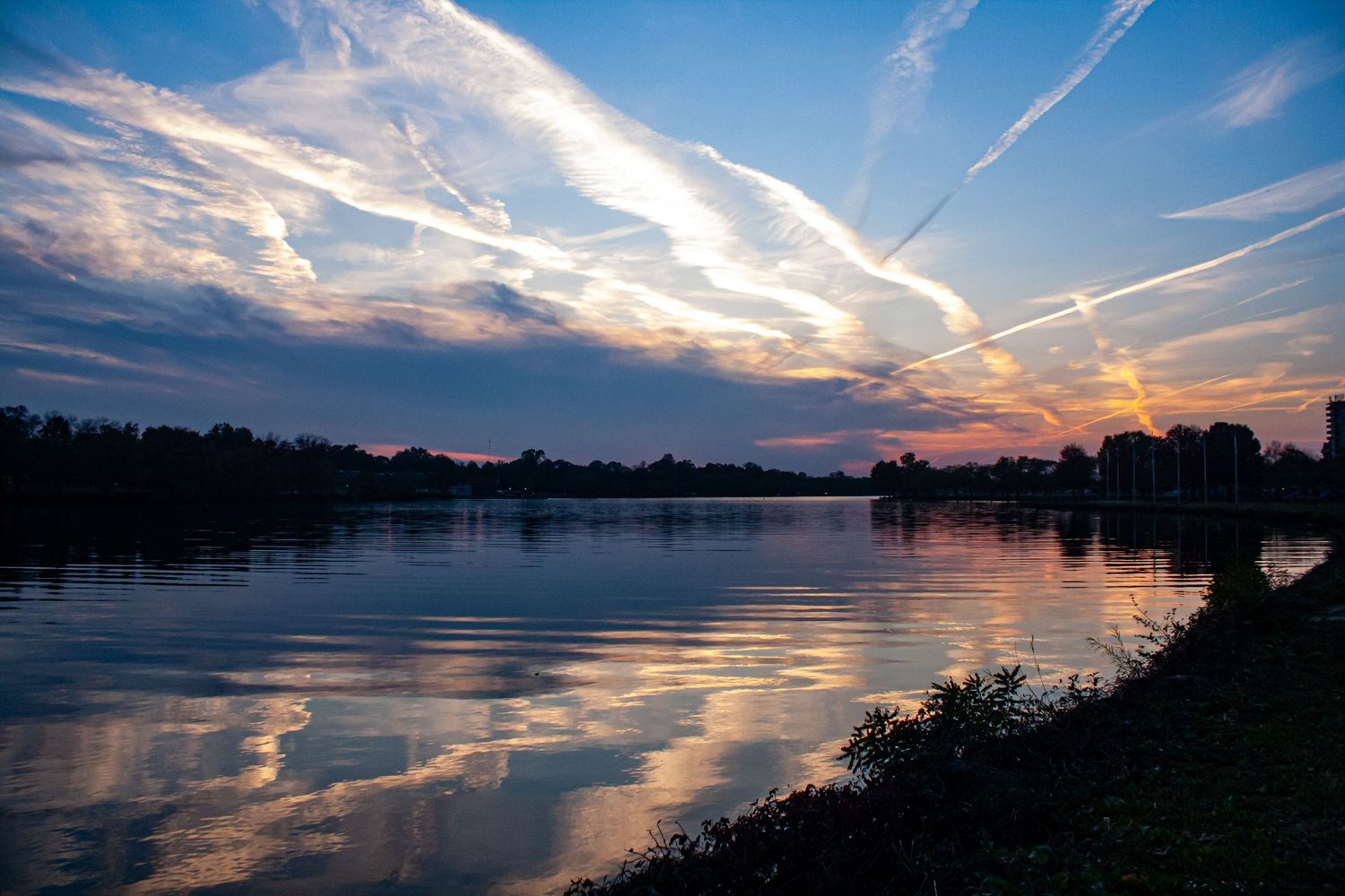 Dramatic sunset sky with streaking clouds reflects on calm lake waters as silhouetted trees line the shoreline at dusk.