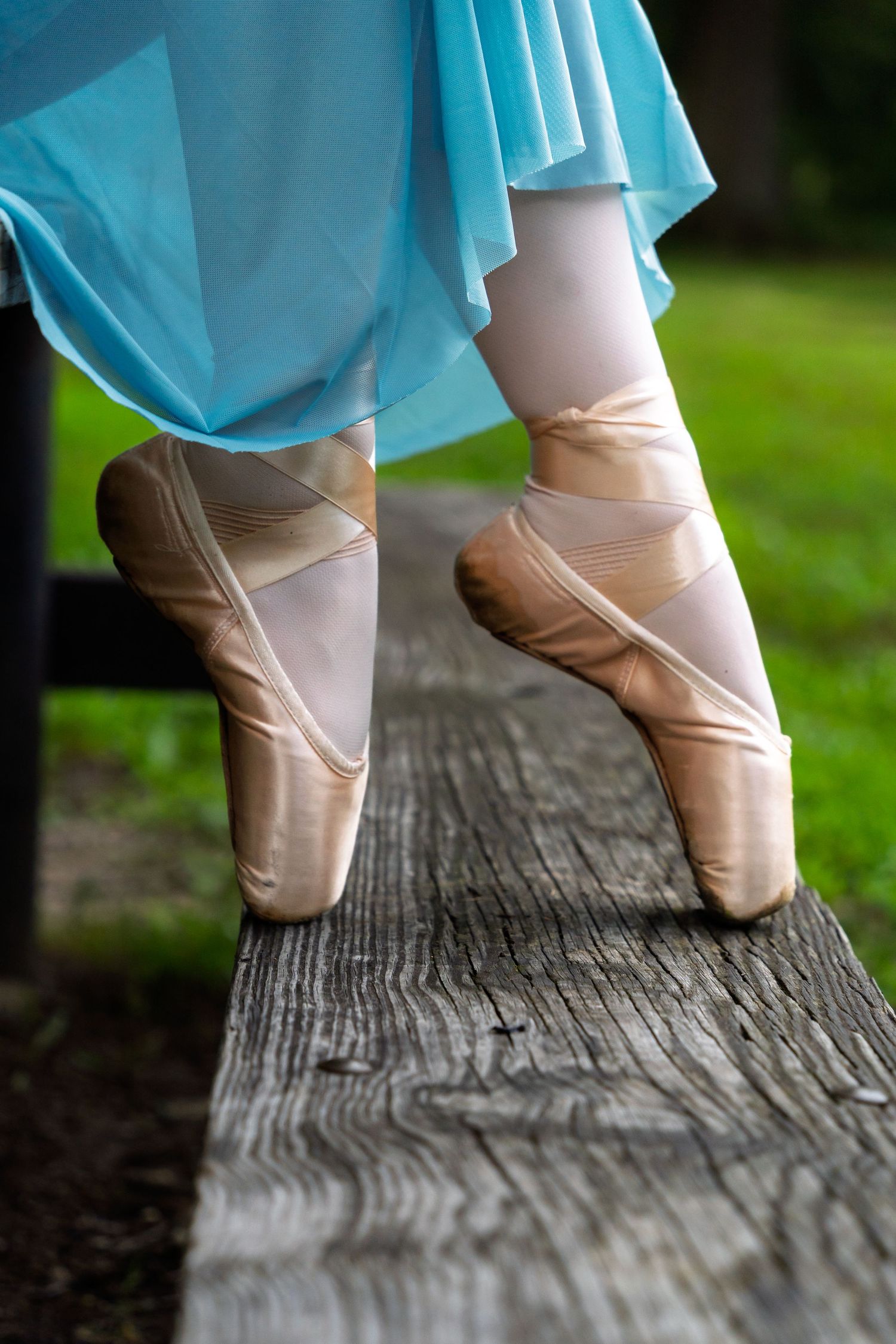 Ballet dancer wearing pink pointe shoes and blue skirt balances on weathered wooden beam outdoors.