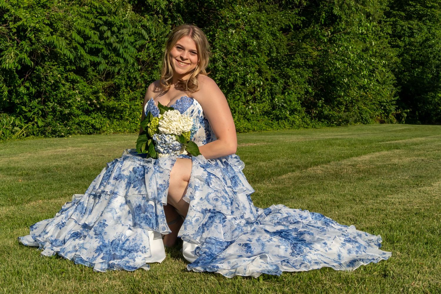 Young woman in blue floral ball gown holding white bouquet, posing outdoors on green lawn for prom photos.