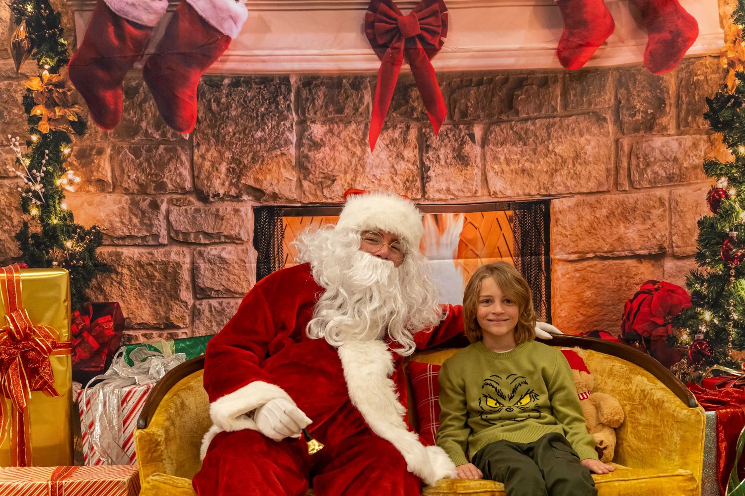 Santa and a visitor in green share a festive moment on a yellow couch by a decorated stone wall.
