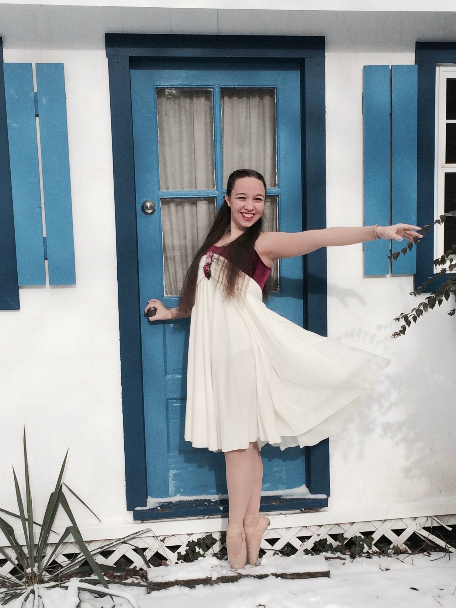 A dancer in a white flowing dress poses against a white building with blue shutters.