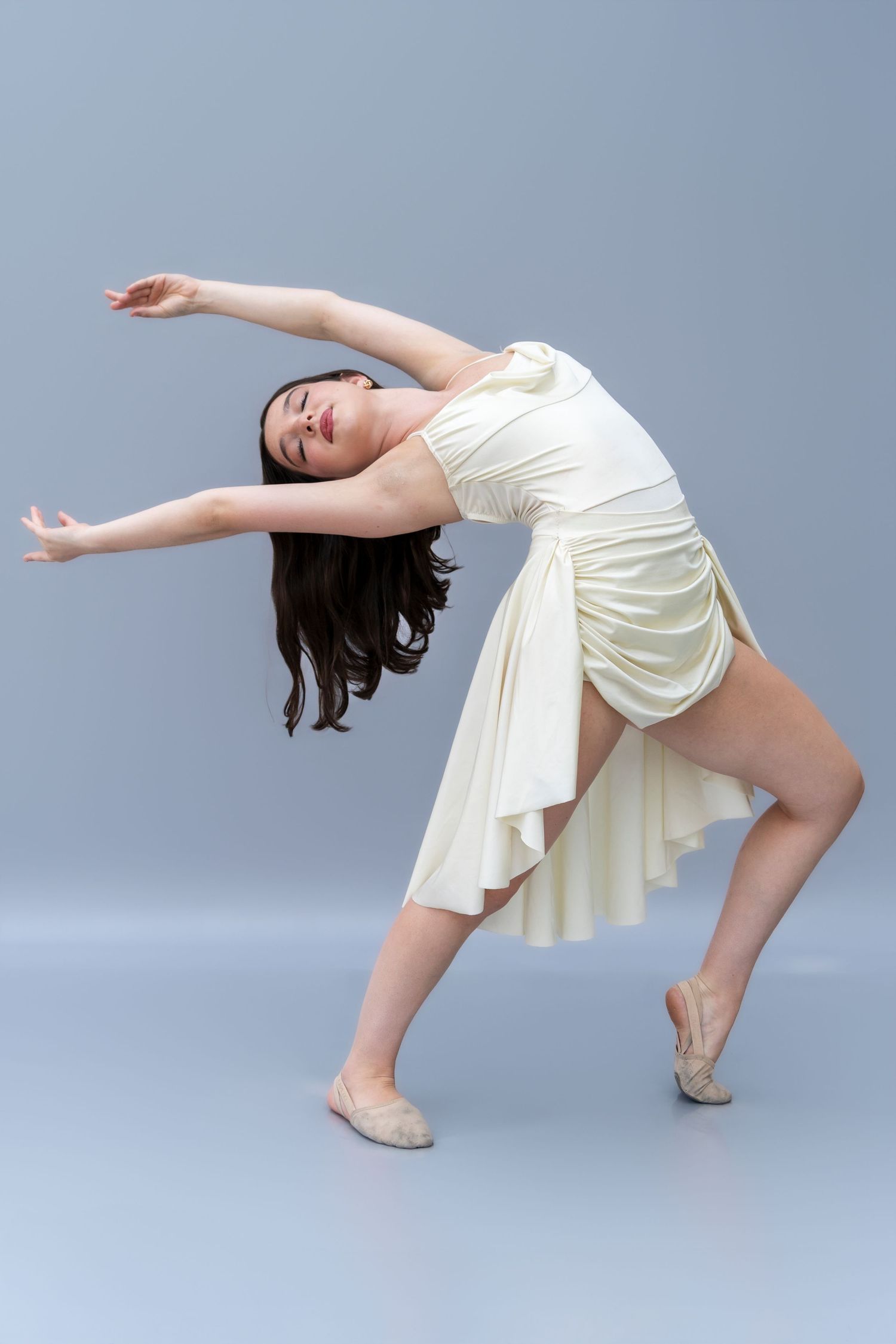 Dancer in flowing white dress performs graceful backbend movement against blue studio background.
