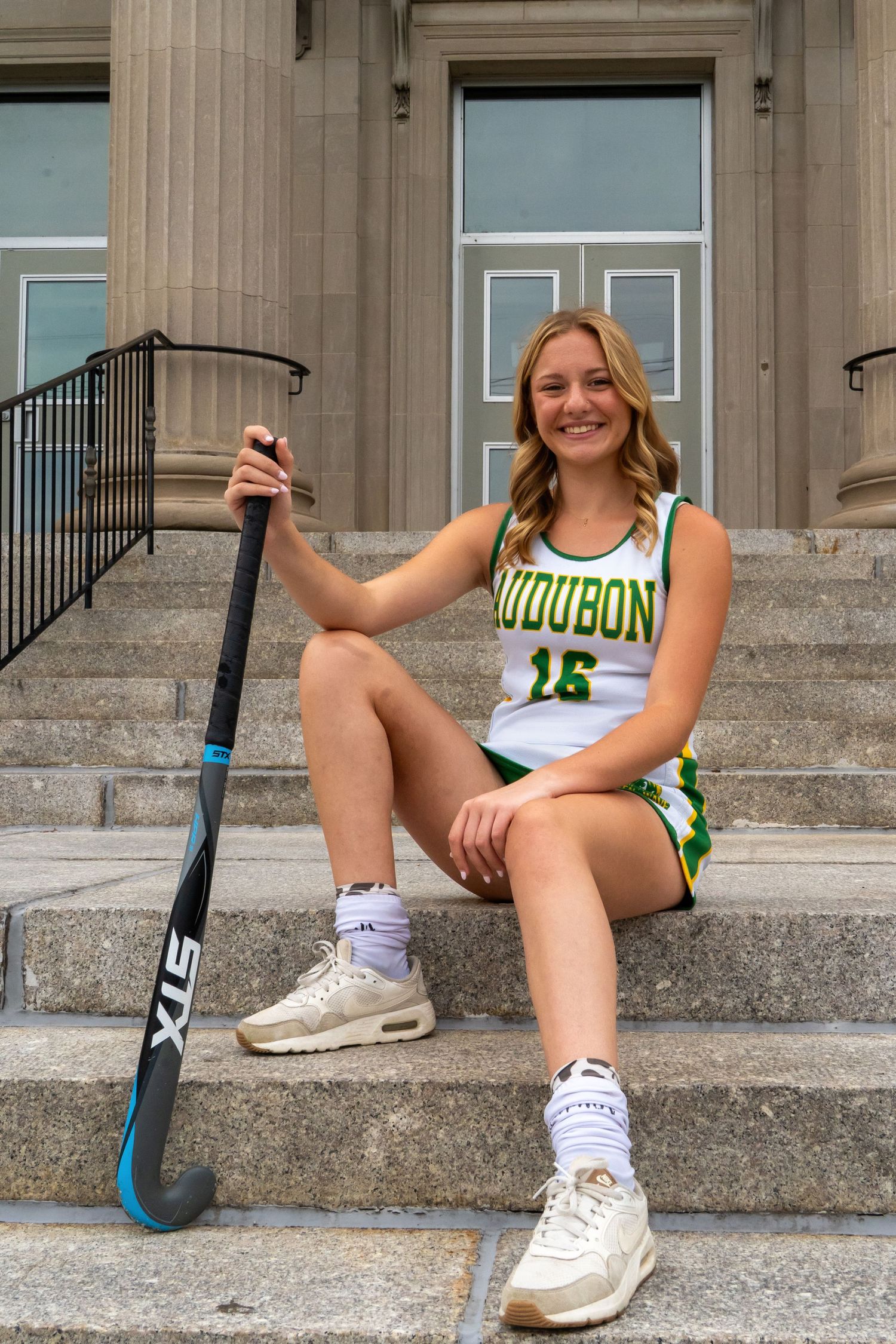 Field hockey player in Audubon uniform sits on stone steps with stick and athletic gear.