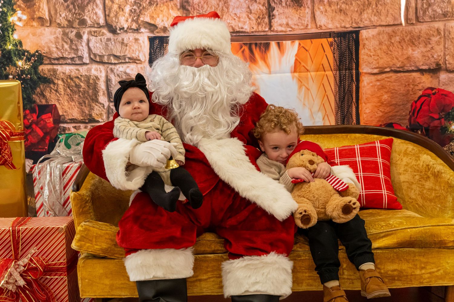 Santa sits with two young children and teddy bears on a yellow couch in front of a stone fireplace.