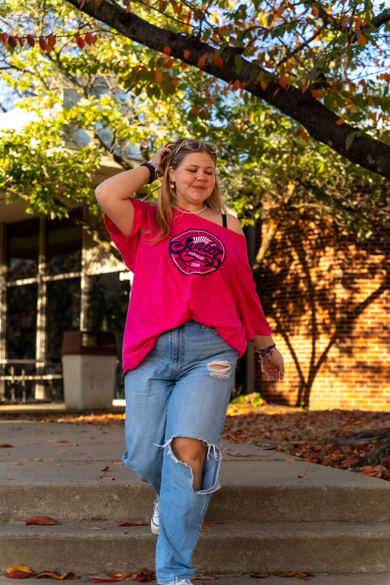 Person in pink shirt and jeans poses playfully in autumn setting with brick building in background.
