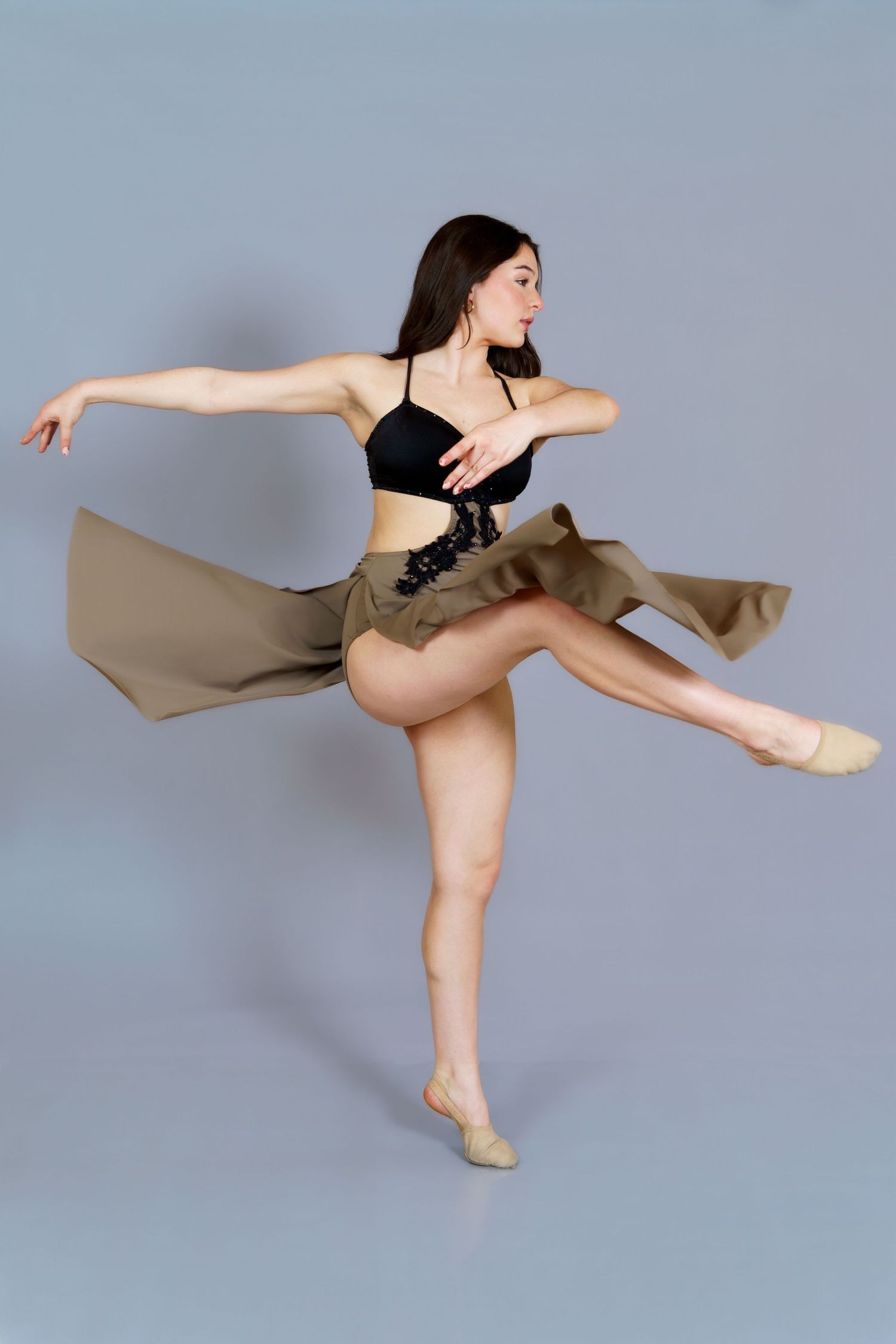 A dancer in a black crop top and flowing olive skirt balances on one leg in a graceful contemporary dance pose.