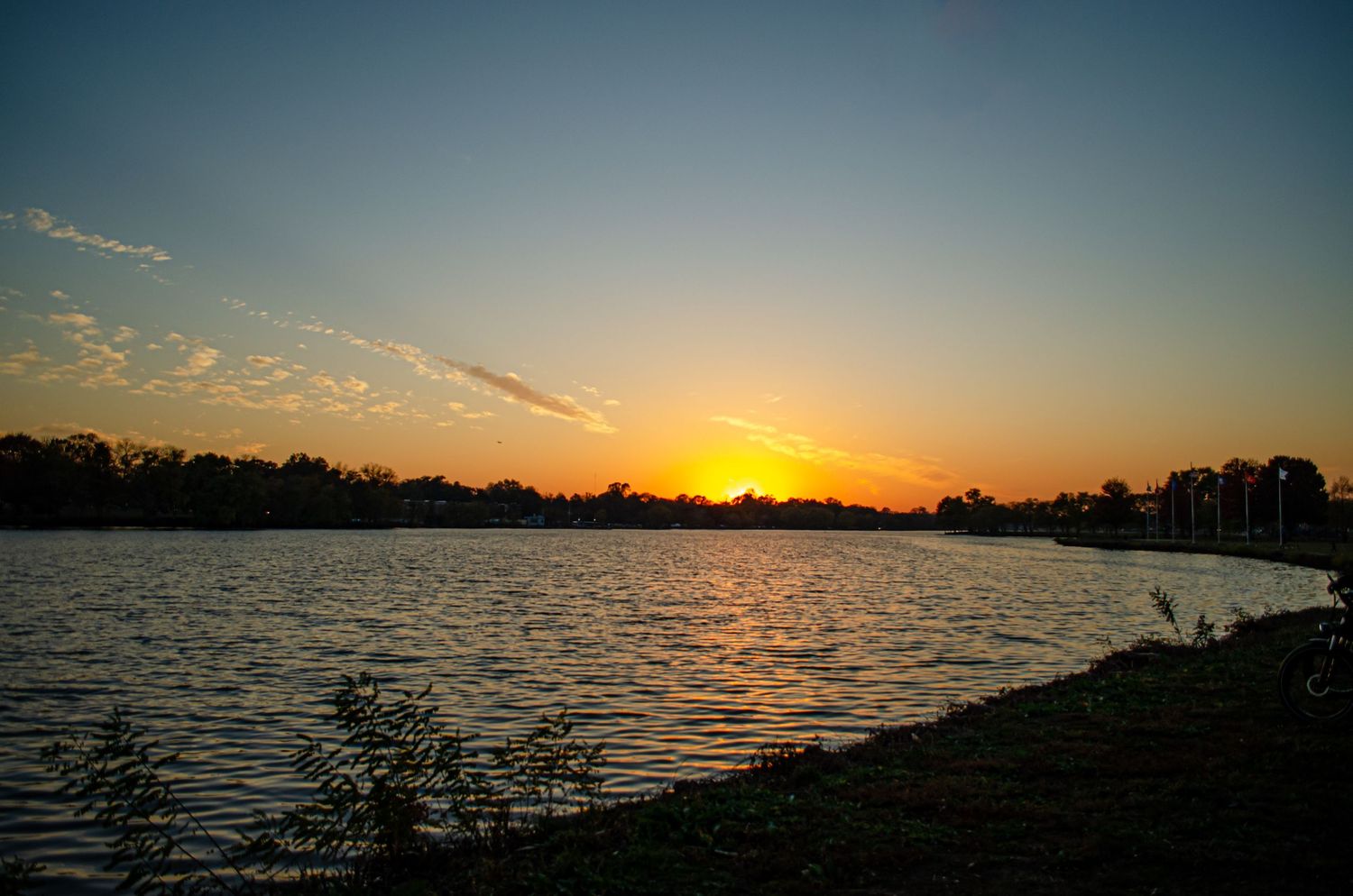 Golden sunset illuminates a tranquil lake with silhouetted trees along the shoreline and wispy clouds in the sky.