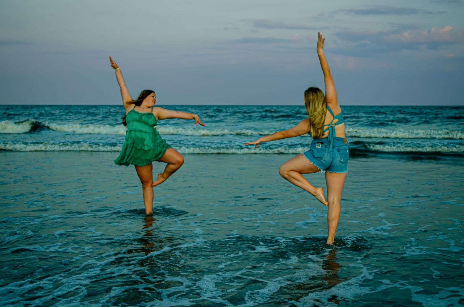 Two dancers in matching teal dresses perform synchronized poses at sunset on the beach.