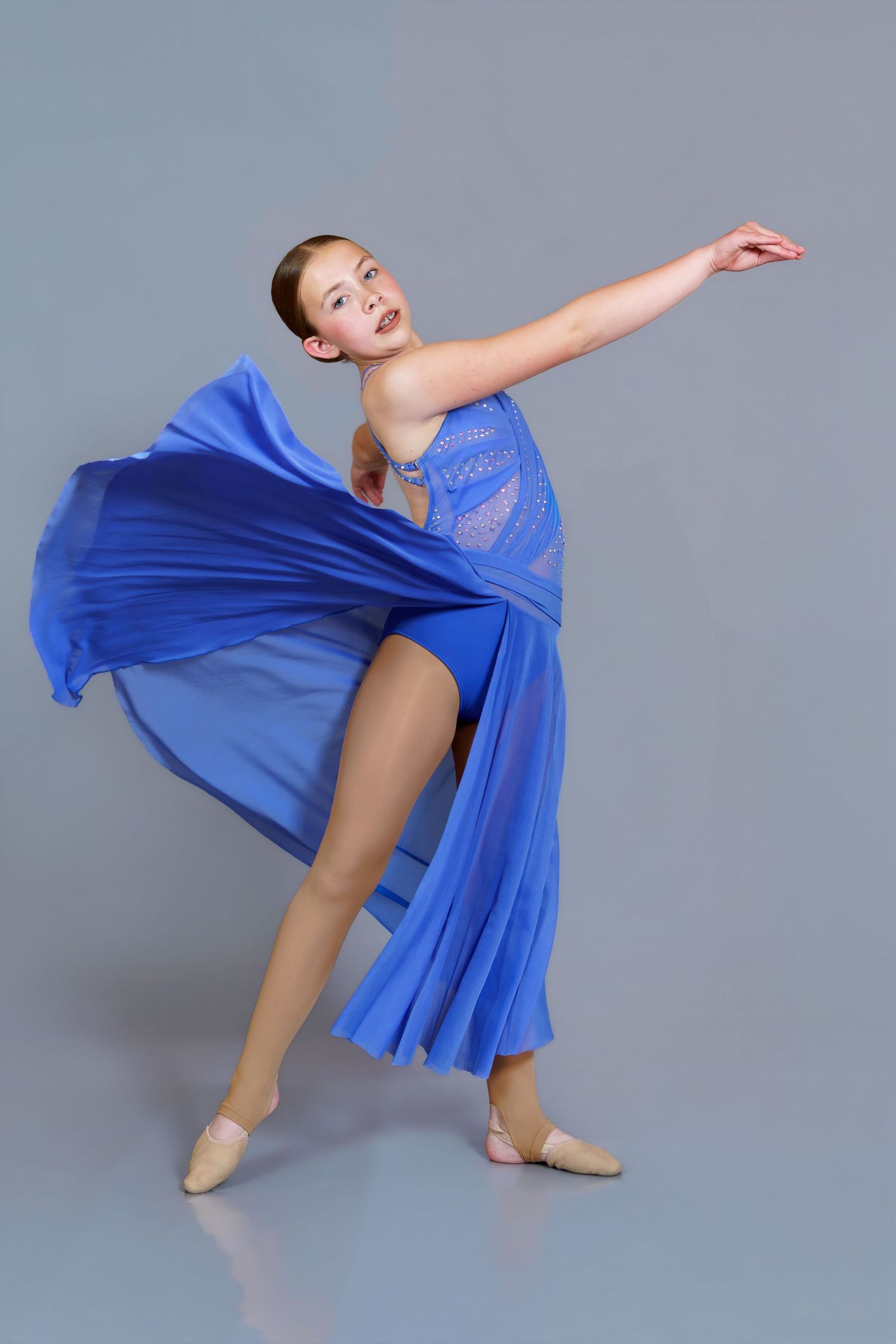 Graceful dancer in flowing blue dress strikes an elegant pose with arm extended in a studio.