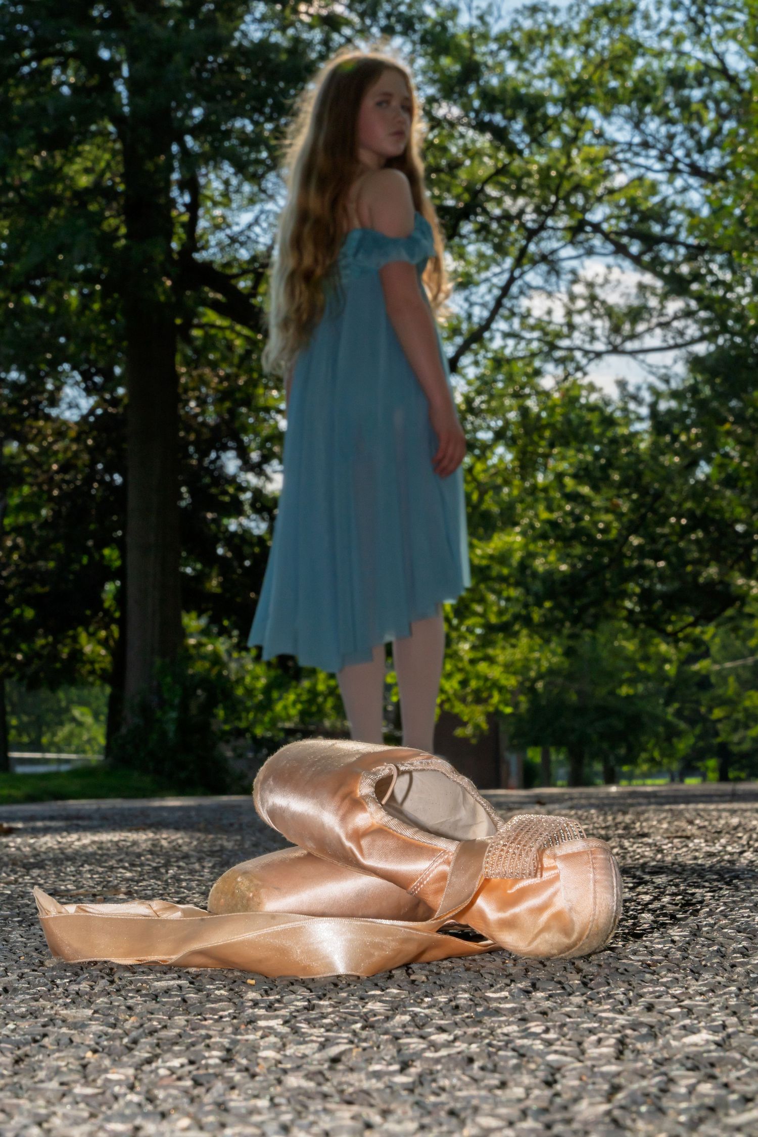 A pair of pink ballet shoes on pavement with a figure in a flowing blue dress standing in sunlit background.