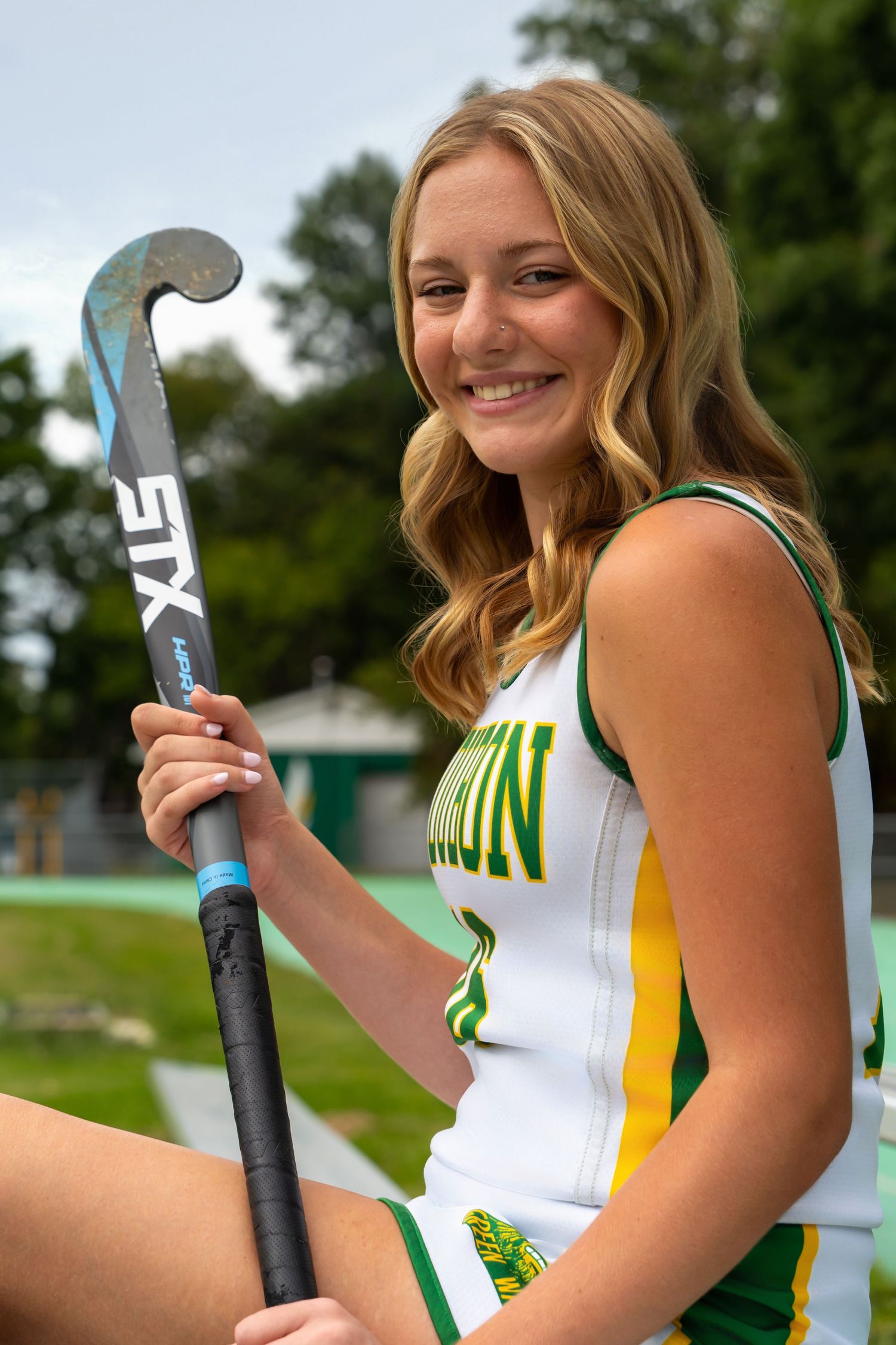 Field hockey player in white and green uniform poses with stick against blurred outdoor background.