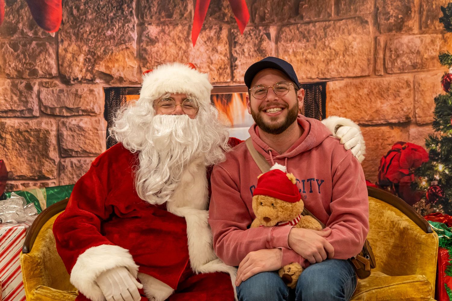 A warm holiday scene with Santa seated next to a visitor holding a teddy bear by the fireplace.