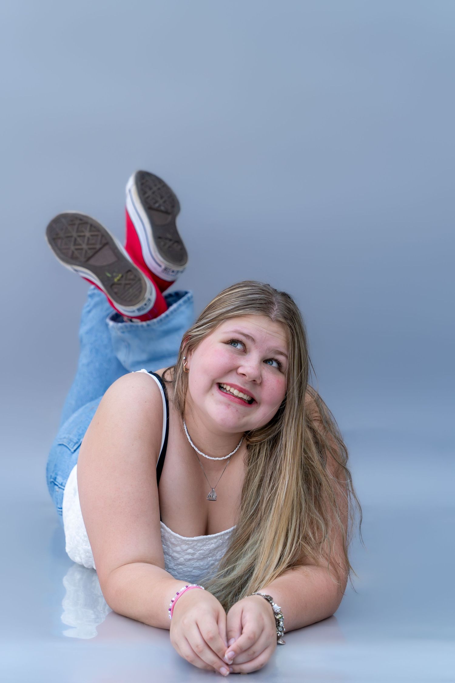 A person in jeans and white top lies on their side in a casual photo studio pose against a gray backdrop.