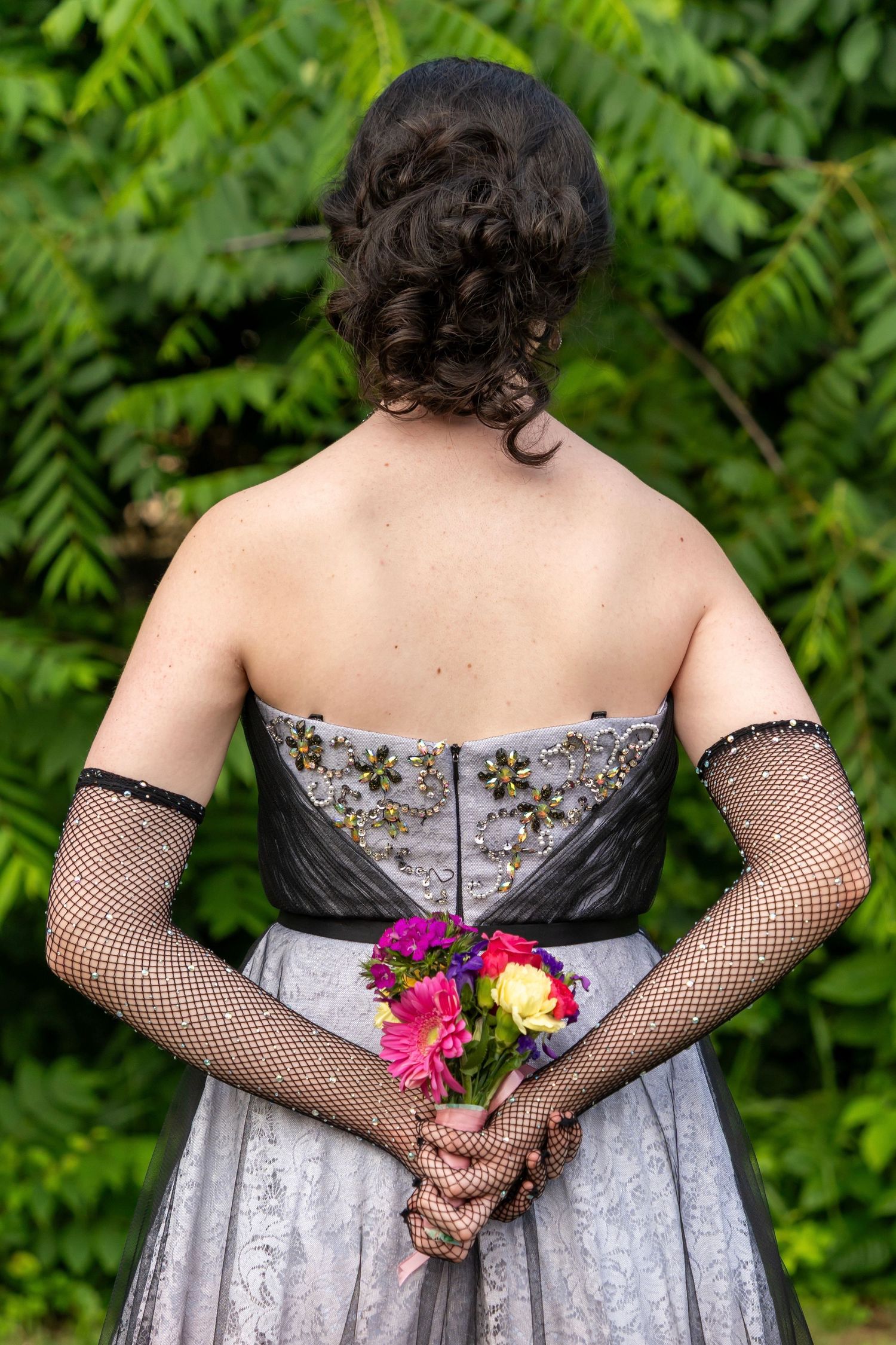 Back view of formal dress with fishnet gloves holding colorful bouquet against green foliage.
