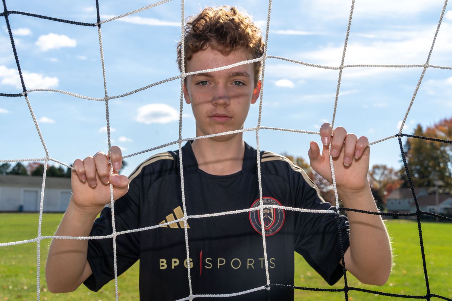 Young soccer player gripping goal net on outdoor field, wearing black BPG Sports jersey on sunny day.