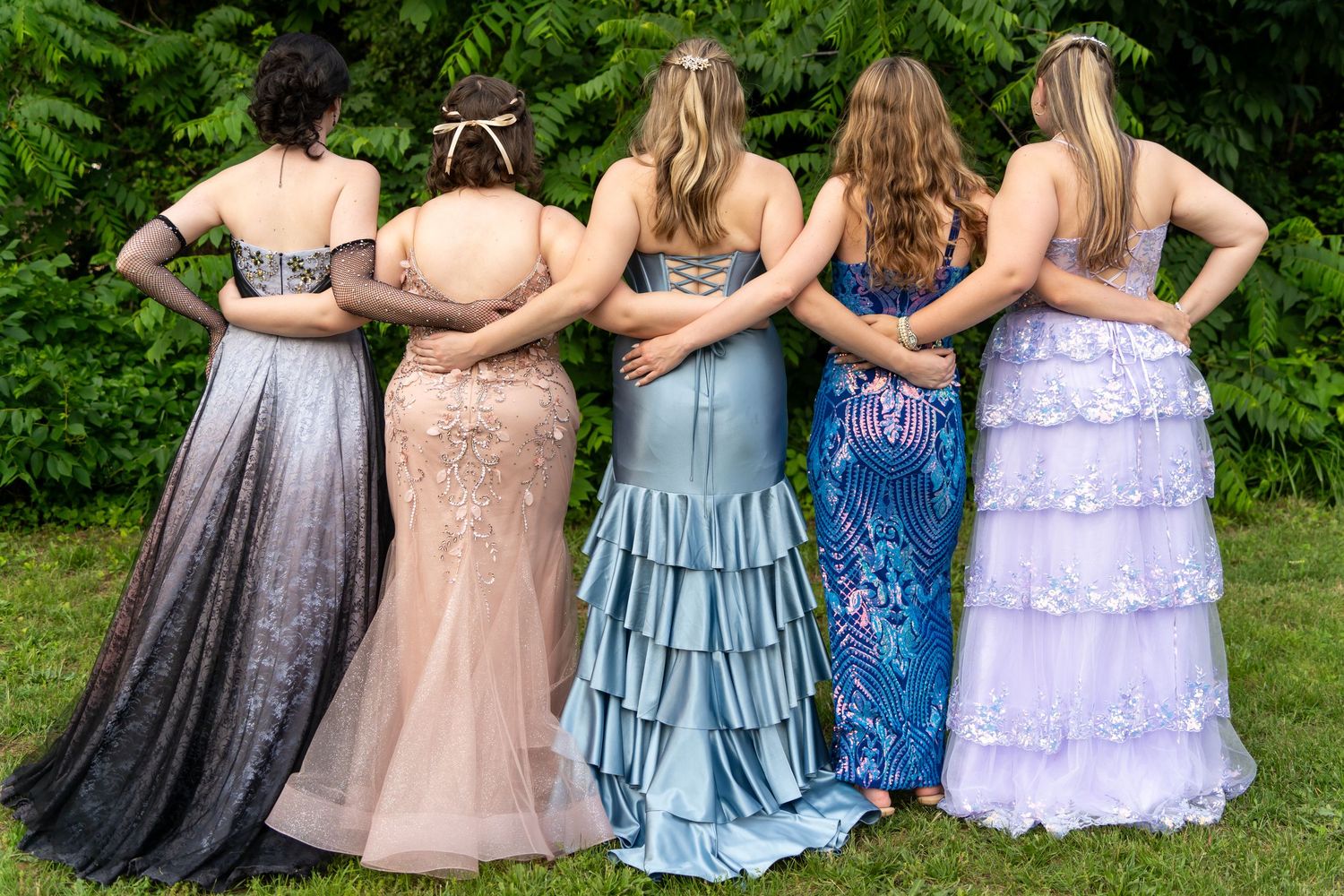 A group photographed from behind wearing elegant prom dresses in grey, blush, blue, floral, and lavender standing arm in arm.