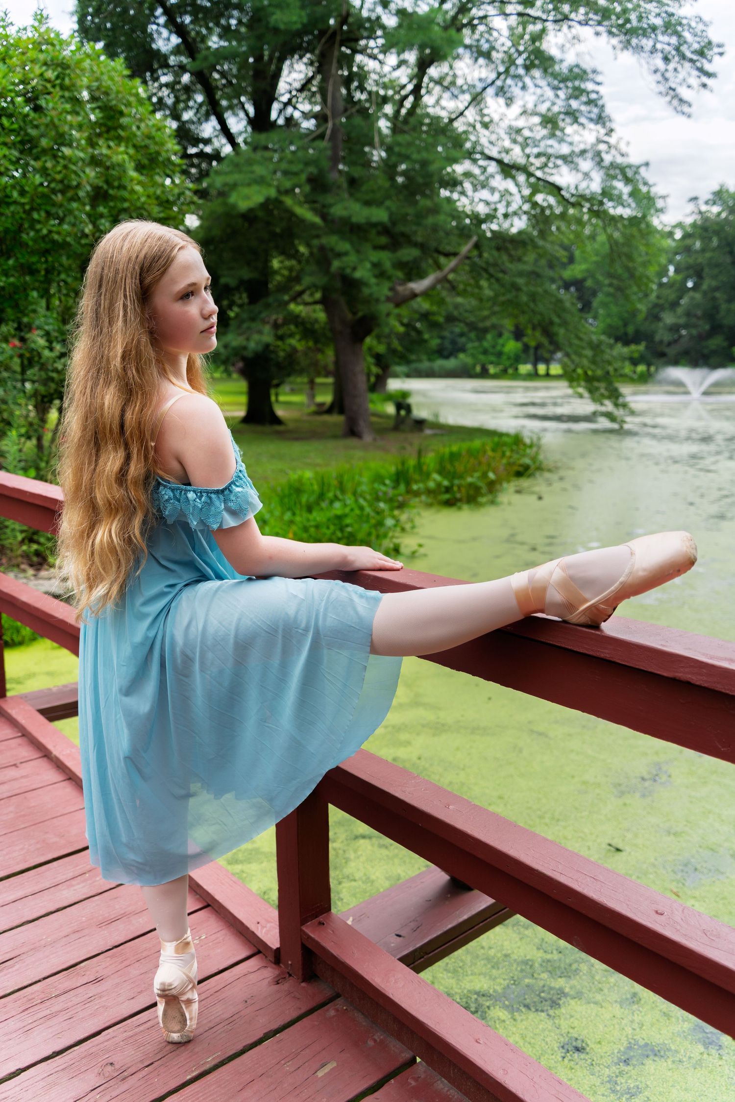 A dancer in a blue dress practices graceful stretches and poses on a wooden deck overlooking a pond.