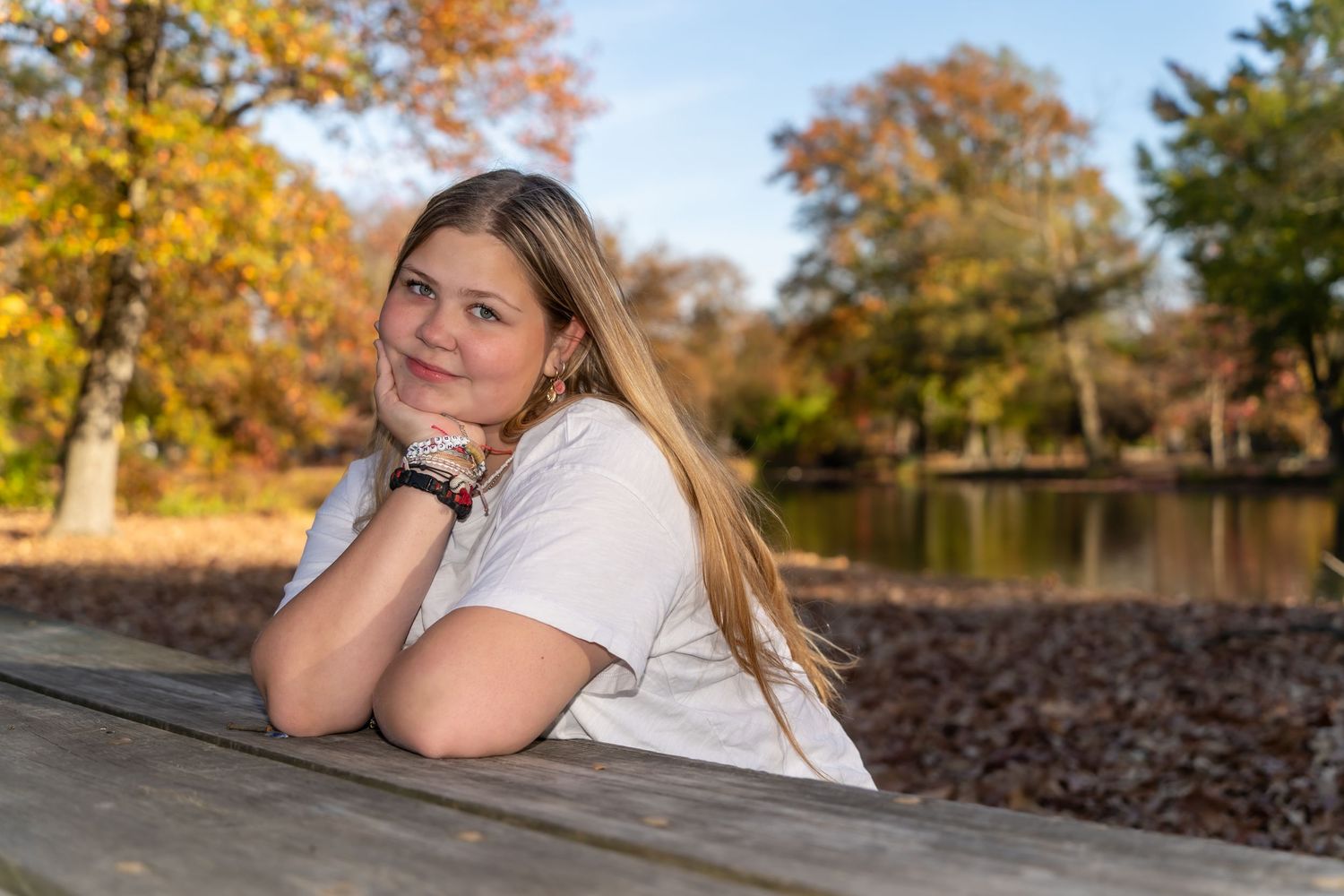 A person in white rests on a picnic table near a pond with autumn trees in the background.