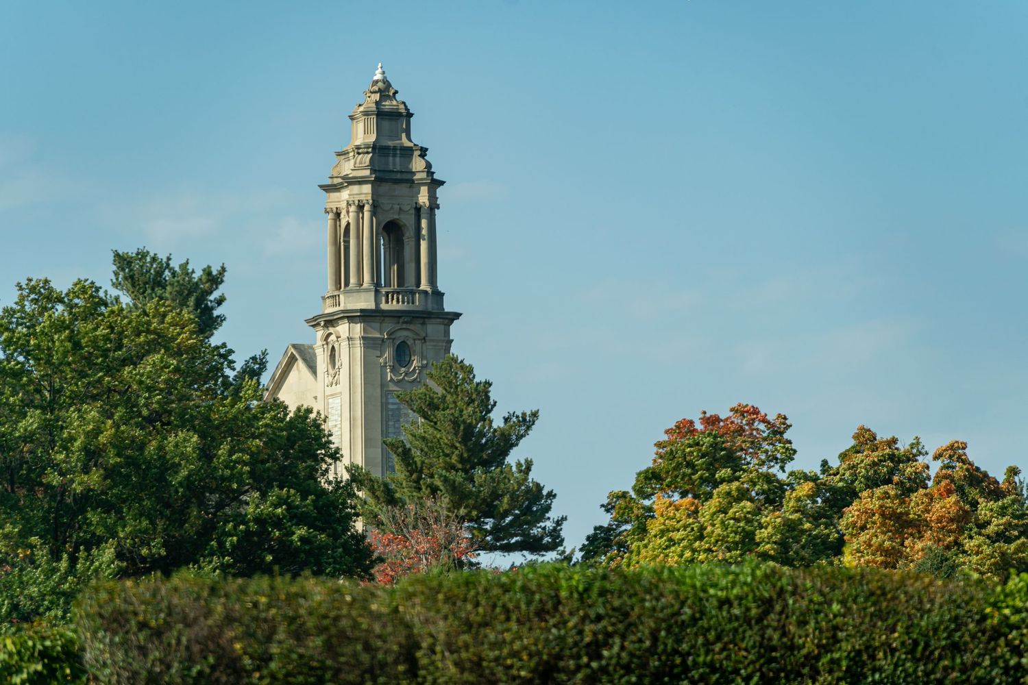 A white stone church tower rises above colorful autumn trees against a bright blue sky.
