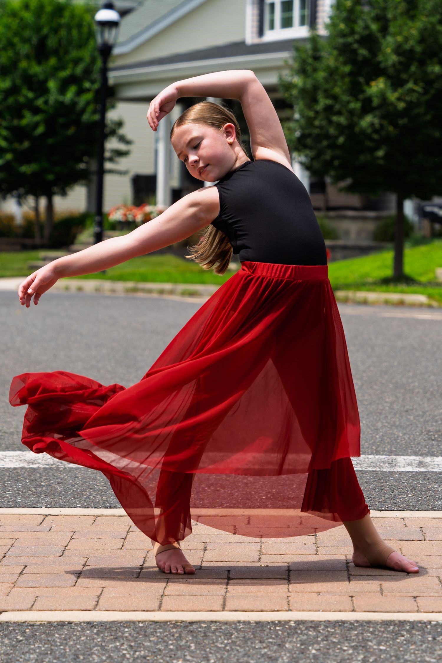 Dancer in a flowing red dress performs an expressive movement on a brick pathway near a grassy area.