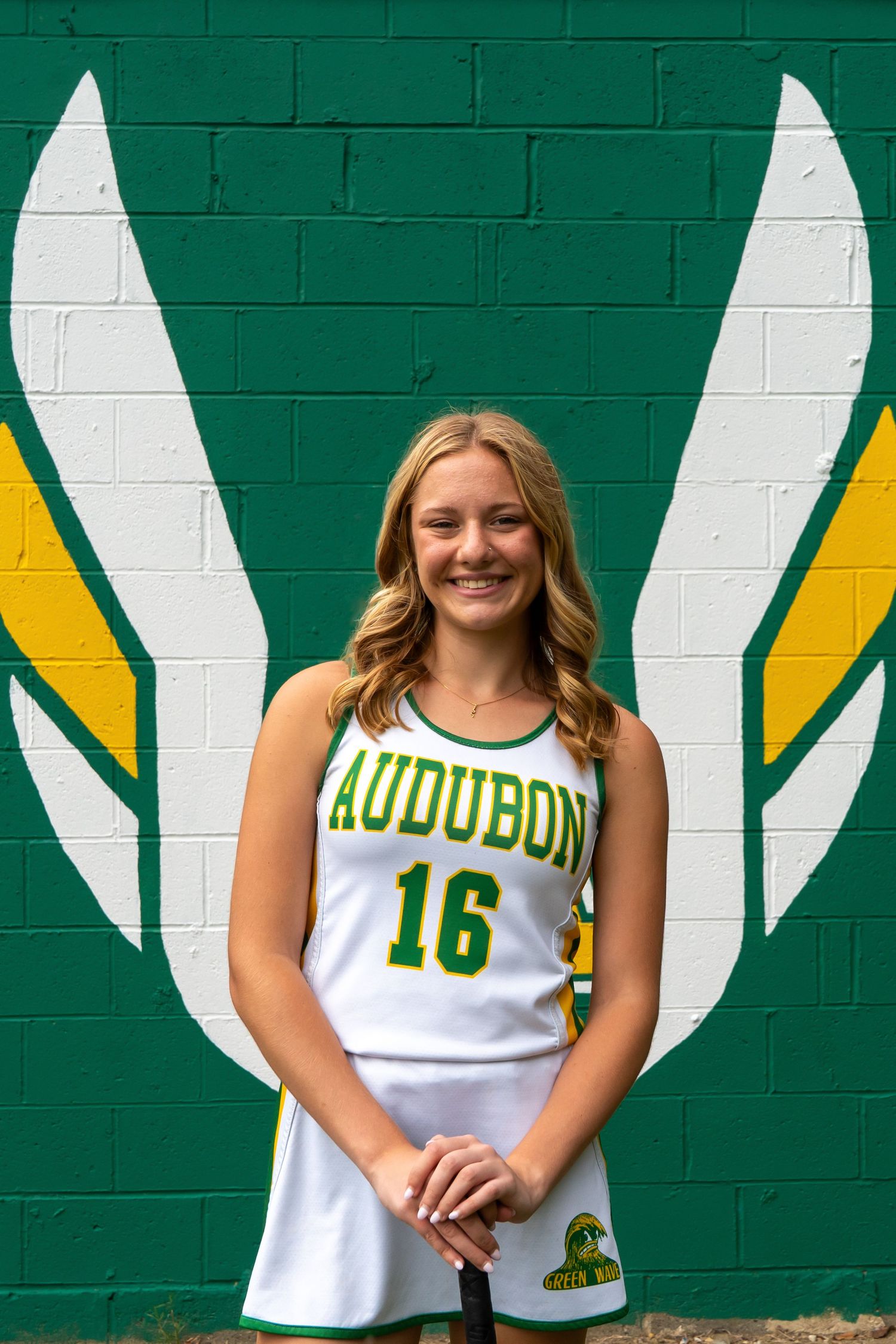 An athlete in a white Audubon #16 uniform stands in front of a green wall with painted white and yellow wing designs.