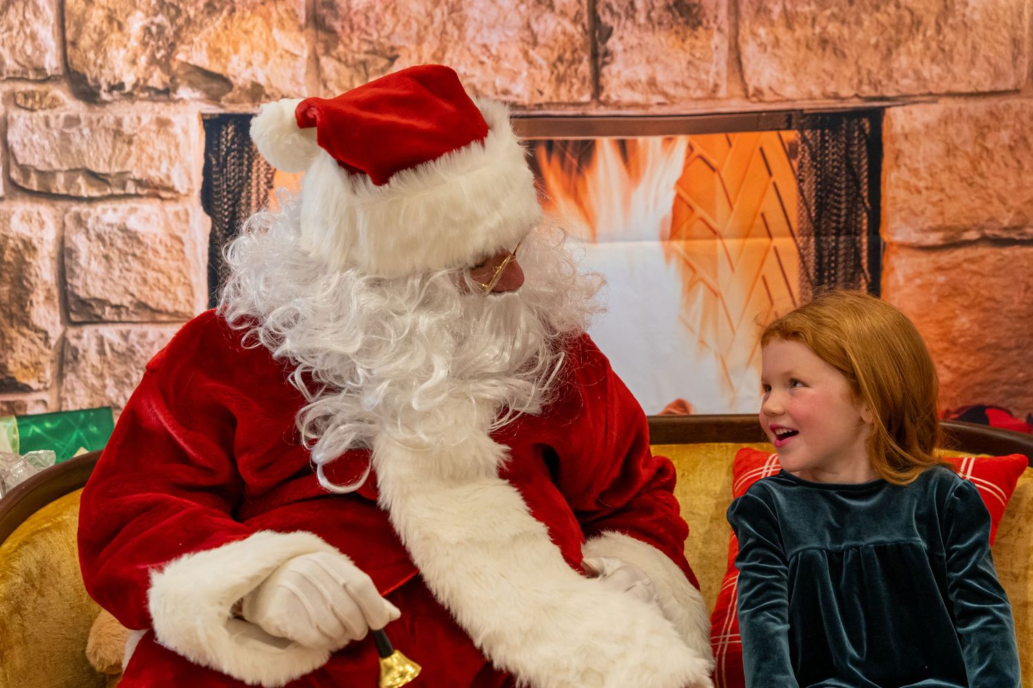 A festive holiday scene with Santa seated on a yellow couch next to a child in green velvet by a stone fireplace.