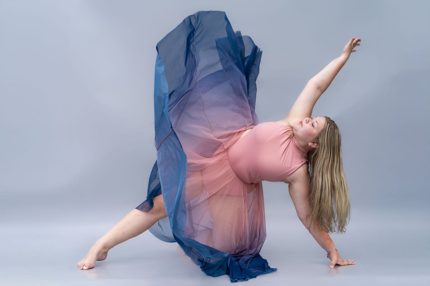 A dancer in a pink leotard and flowing blue fabric performs a graceful side pose against a gray backdrop.