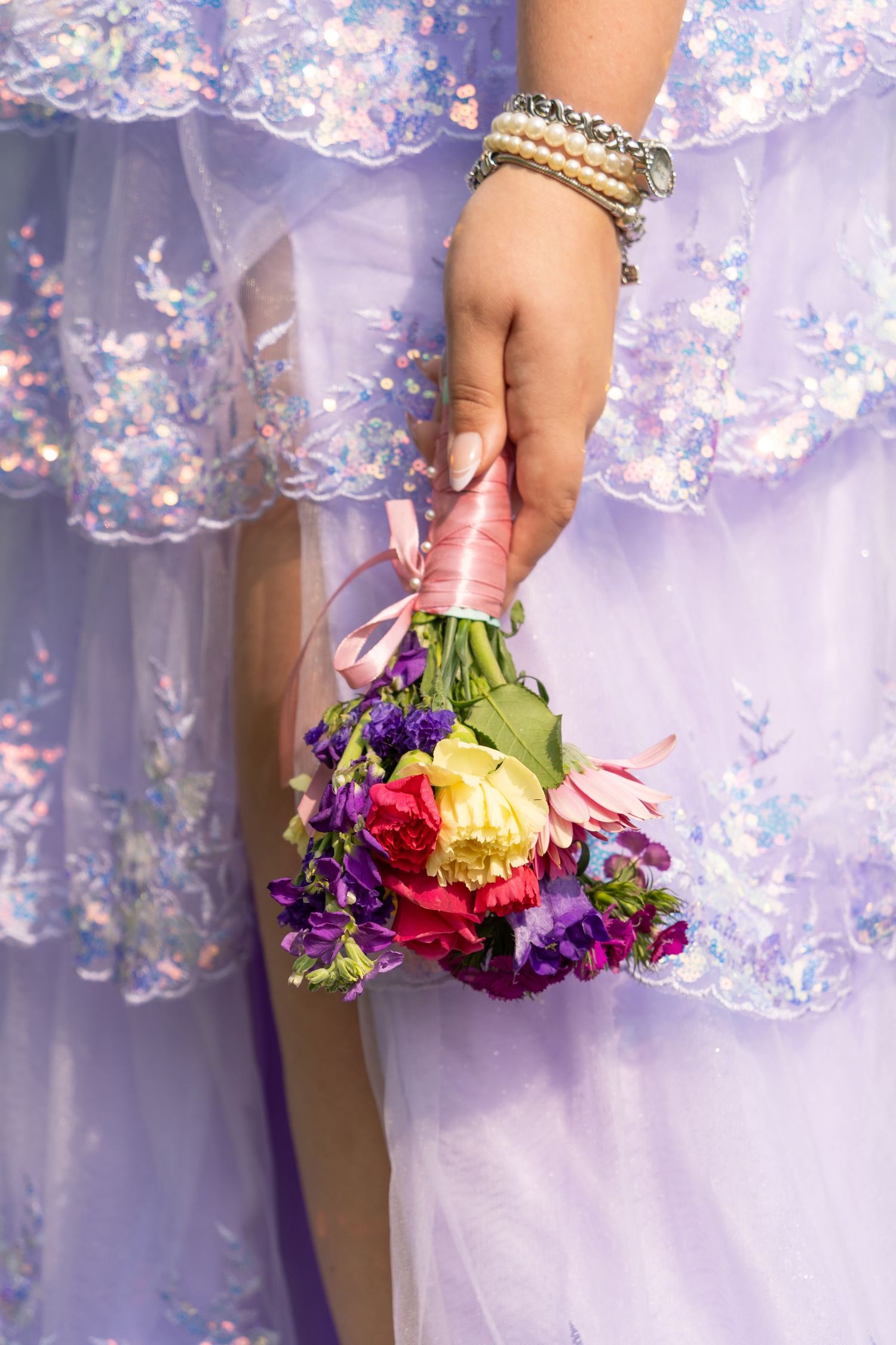 Close-up of hand wearing bracelets holding small colorful bouquet against purple lace prom dress.