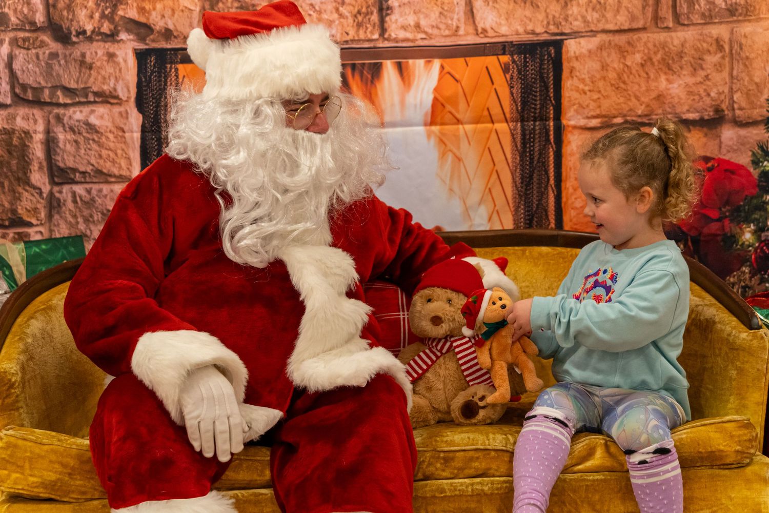 A young visitor in winter attire shares a cheerful moment with Santa on a yellow couch.