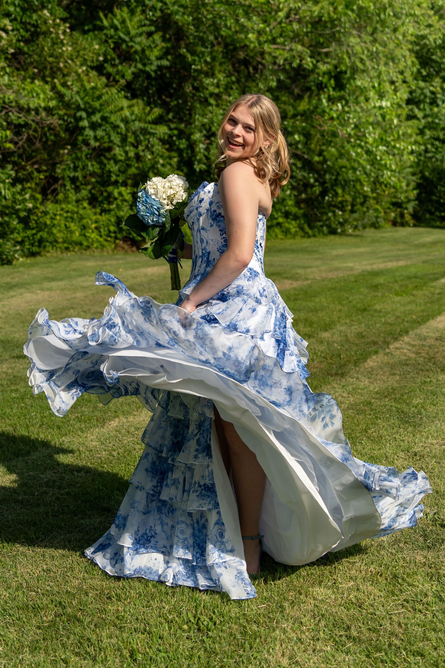 Young woman twirling in a blue floral gown holding white hydrangeas bouquet on a green lawn.