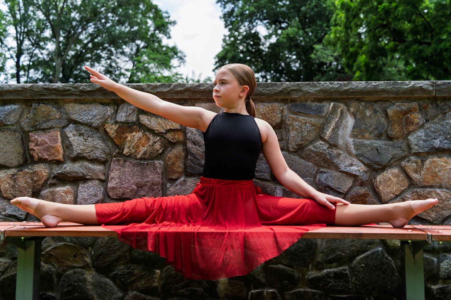 A dancer in a flowing red dress performs graceful movements along a rustic stone wall in the garden.