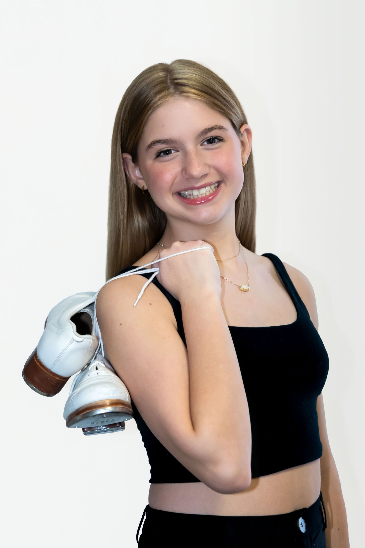 Smiling teenage girl holding white ice skates over shoulder, wearing black crop top and gold necklace, studio portrait.