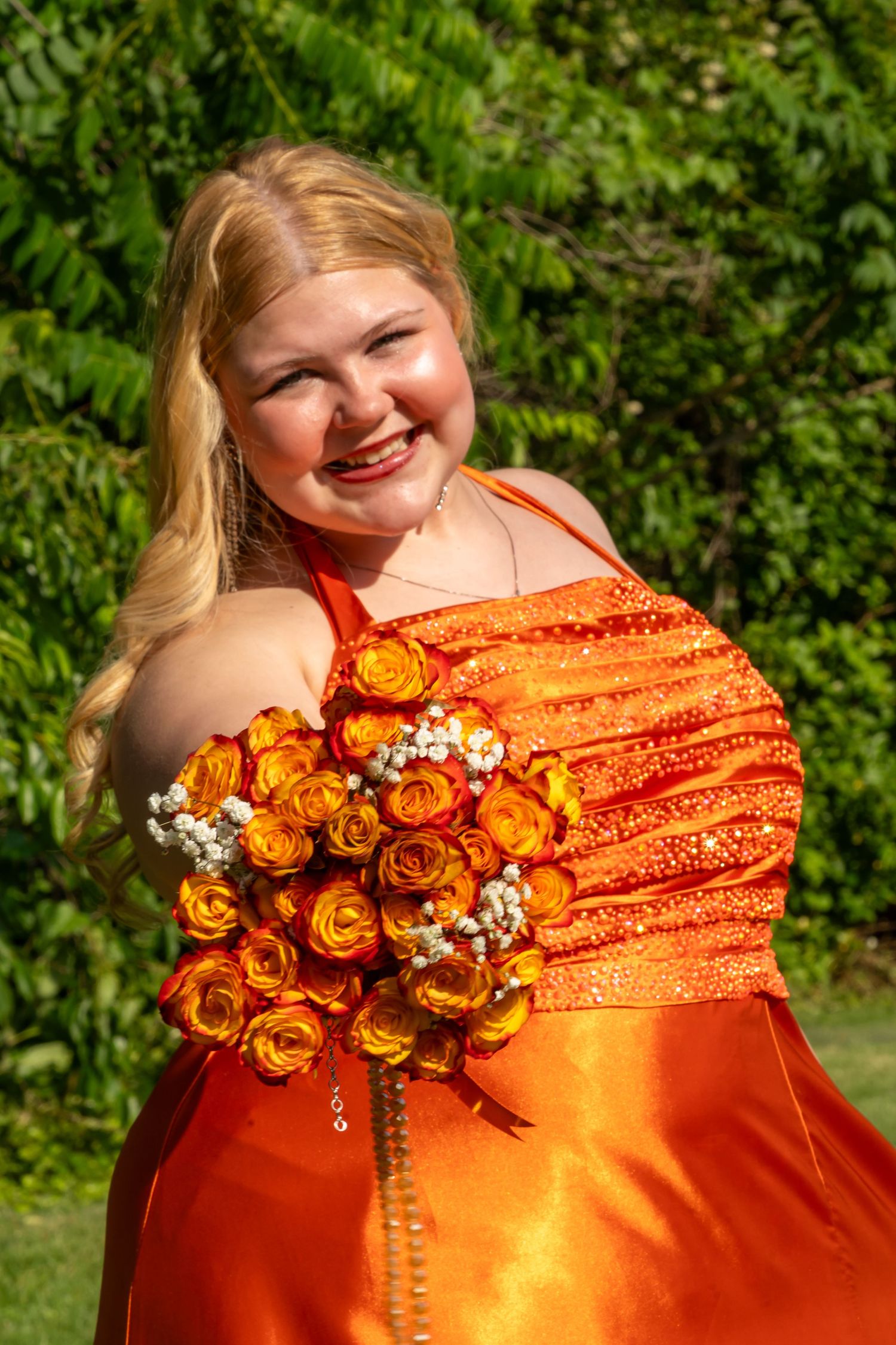 Smiling blonde teen in orange sequin prom dress holding bouquet of orange roses outdoors.