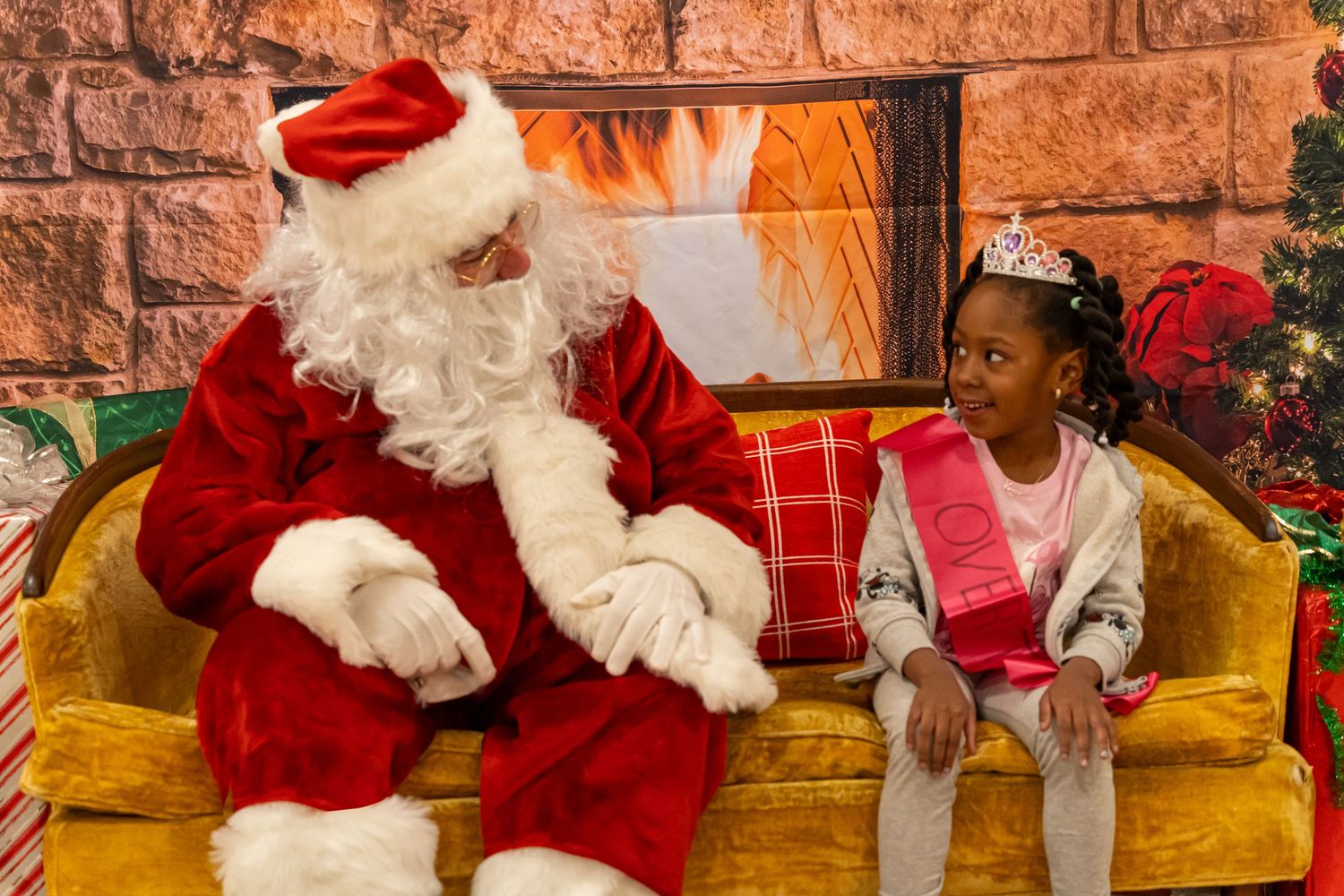 Santa greets a visitor in a festive holiday setting with a decorated stone fireplace backdrop.