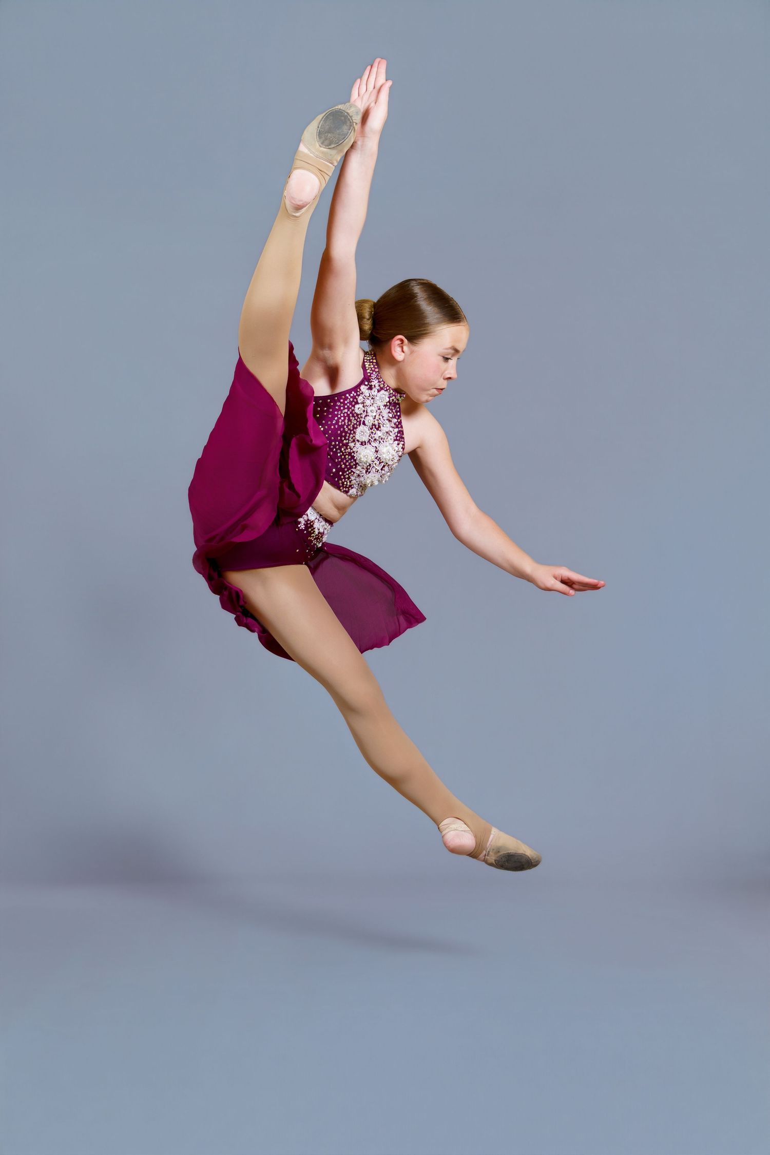 Young dancer in magenta dress performs a stunning leap with leg raised overhead in a studio setting.