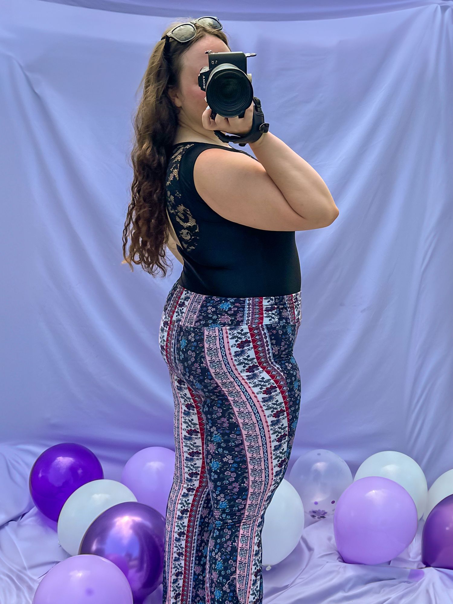 A person in a black tank top and patterned pants poses with a camera against a purple backdrop with colorful balloons.
