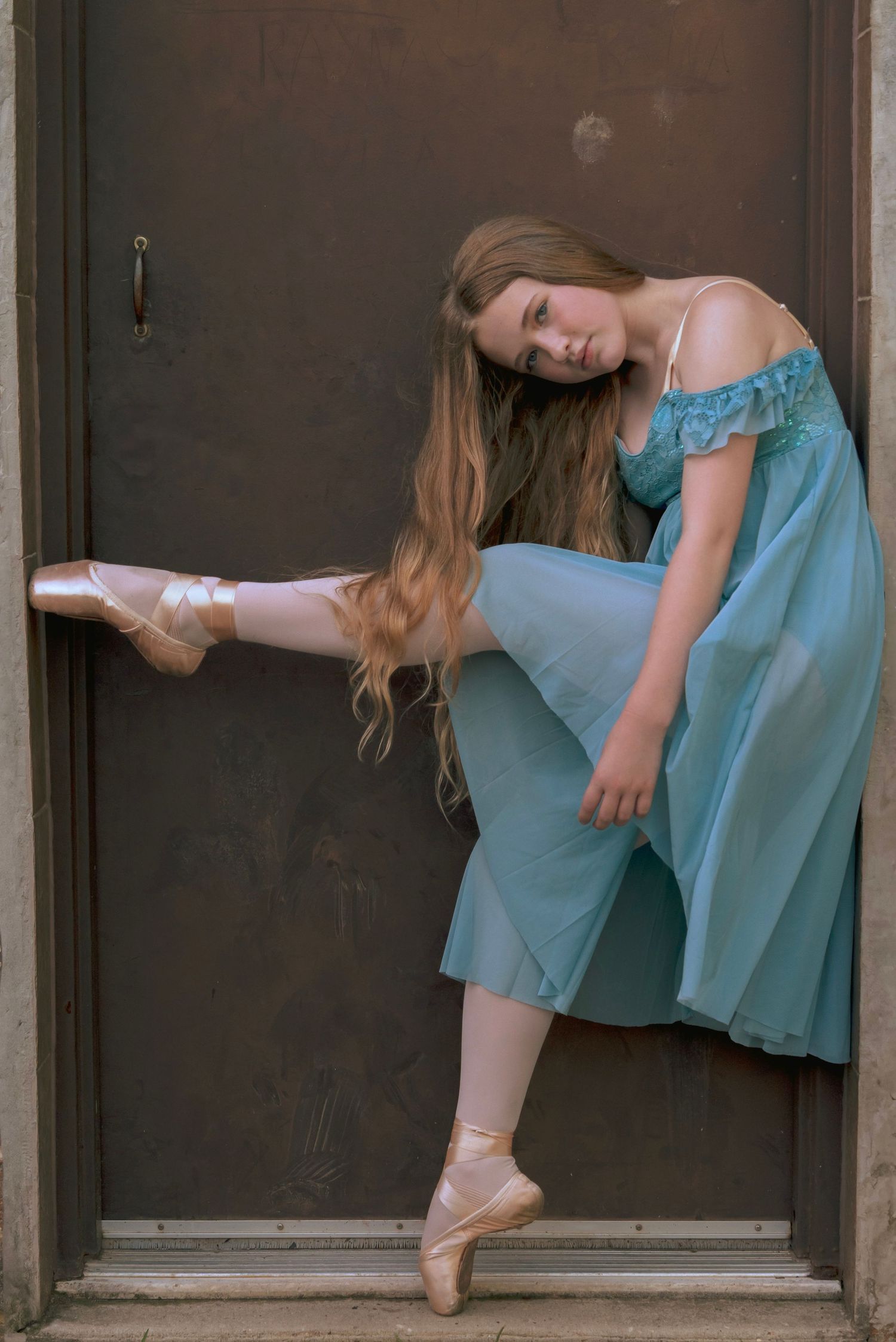 Ballerina in flowing blue dress poses gracefully en pointe against a dark doorway showing elegant dance technique.