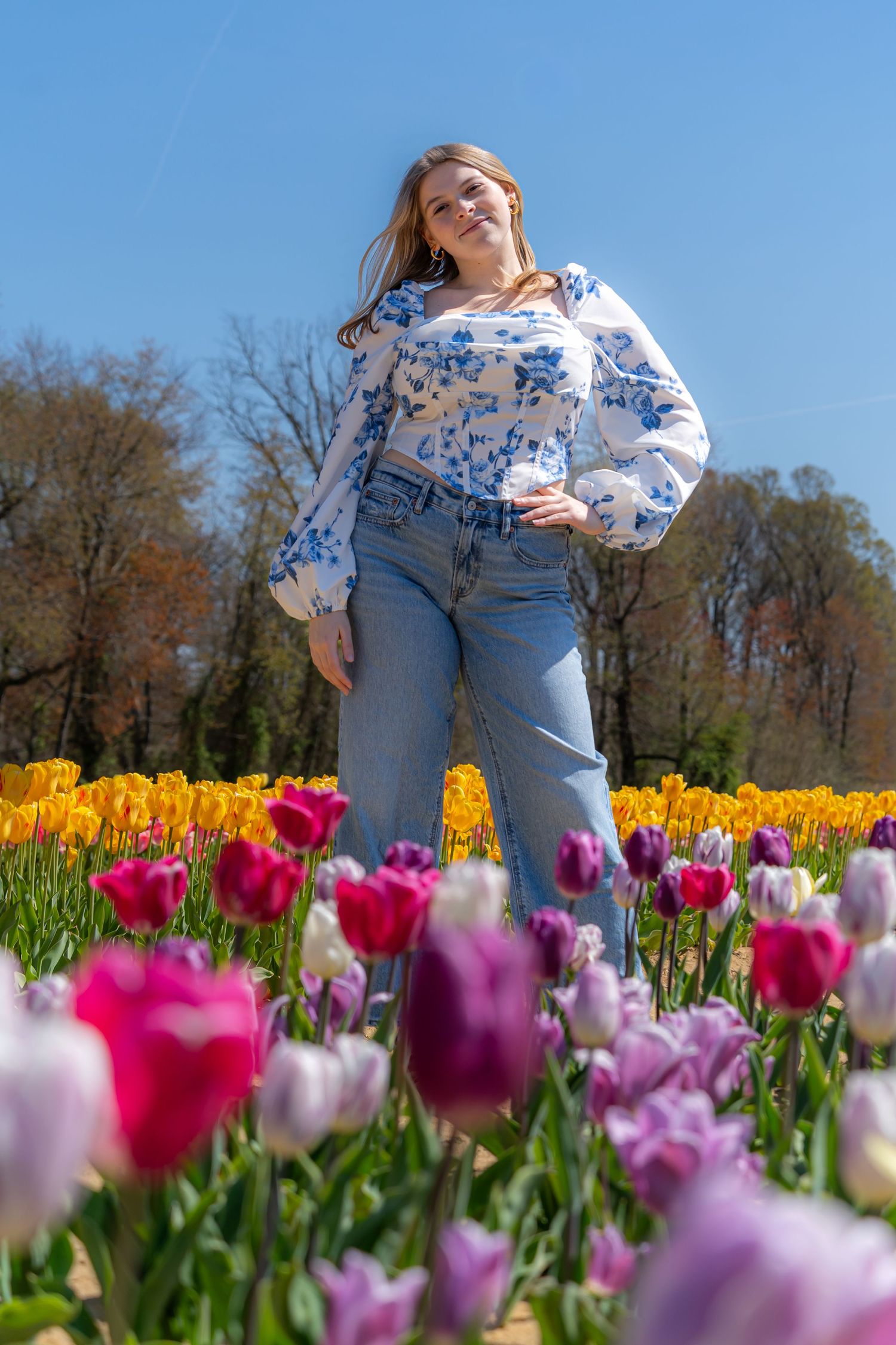 Woman in floral top and jeans posing in a vibrant tulip field with yellow, pink, and purple blooms under blue sky.