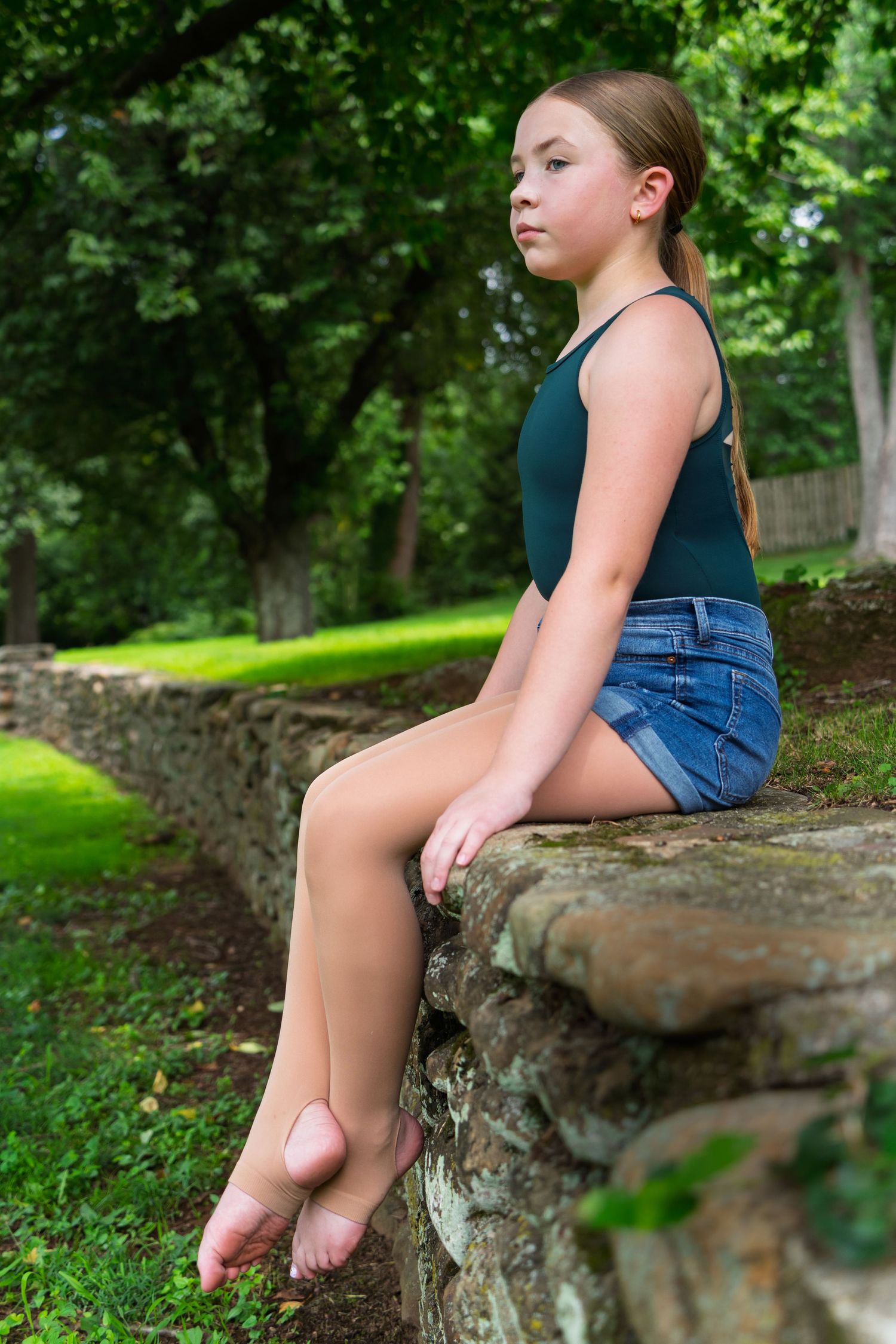 A young girl in denim shorts and dark tank top sits on a stone wall in a peaceful garden setting.