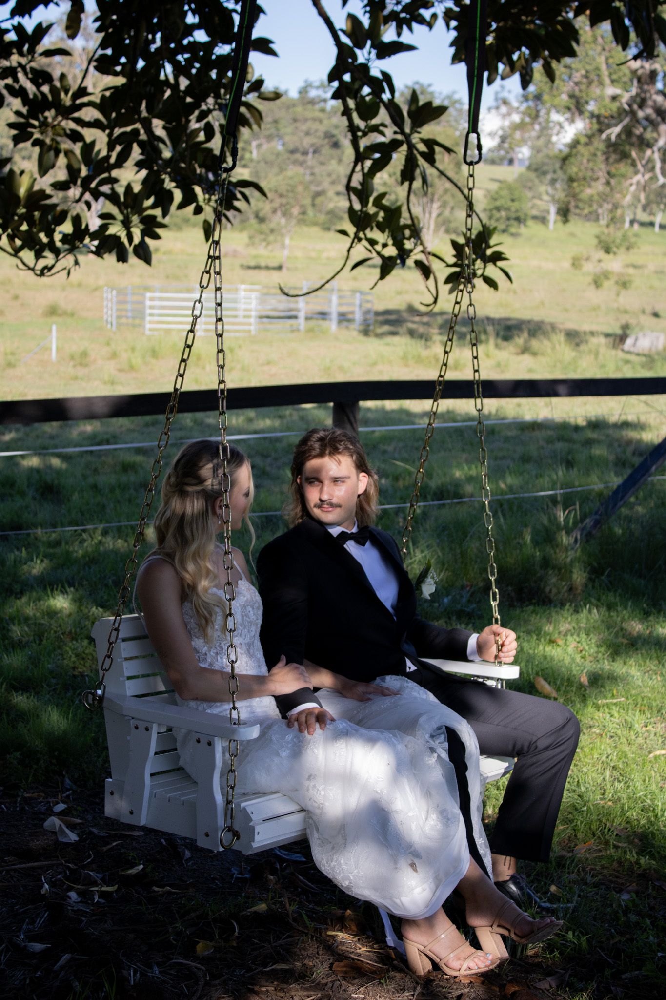 A newlywed couple sitting on a chair swing under a tree.