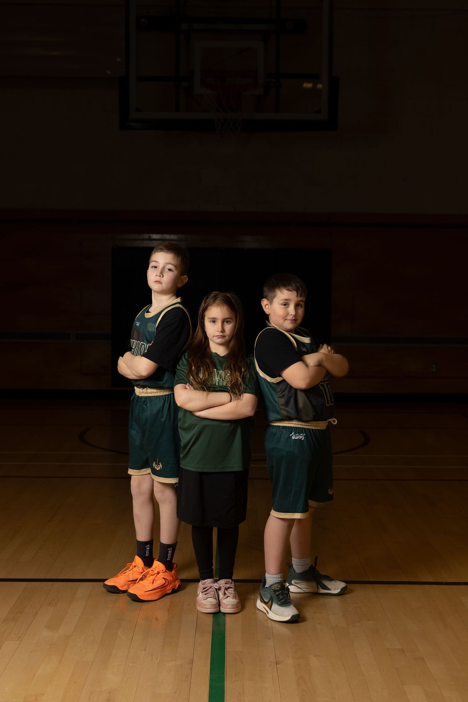 Three children in green basketball uniforms stand with arms crossed on an indoor court, posing as a confident youth team.