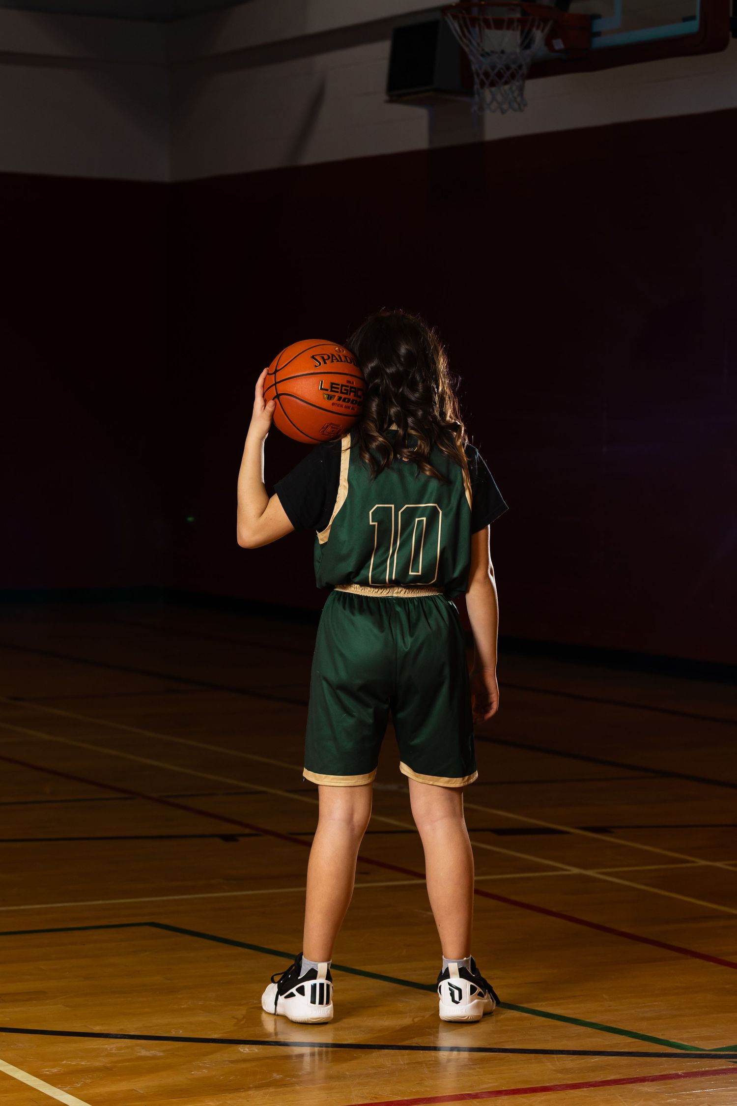 Basketball player in a green number 10 uniform stands on a gym court, back to the camera, holding a basketball up near one shoulder.