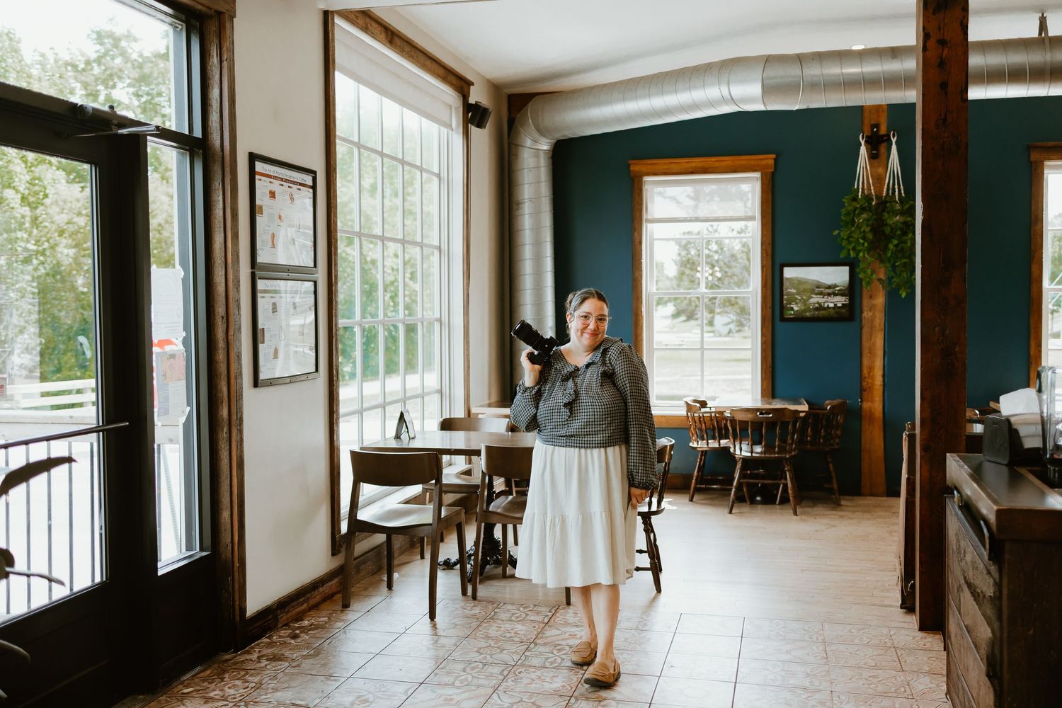 Woman holding a camera stands smiling in a bright rustic cafe interior with wooden tables, large windows and teal walls.