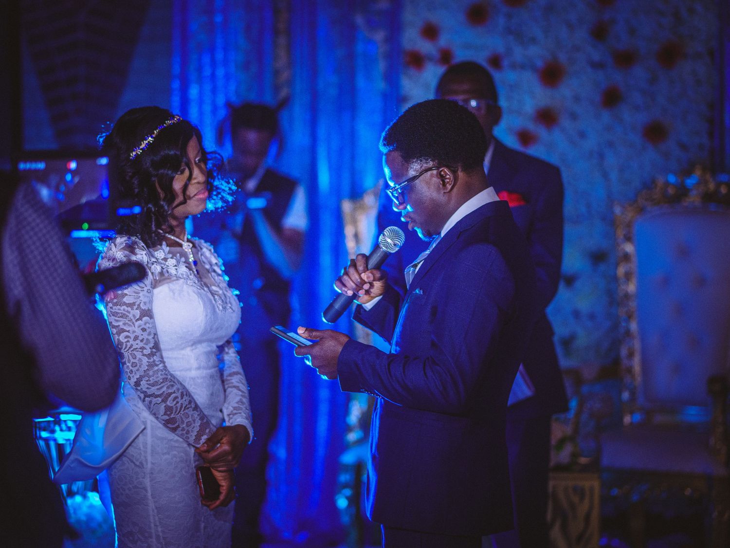 The image depicts a wedding ceremony with a bride in a white lace dress and a groom in a blue suit, exchanging vows with a microphone. Blue